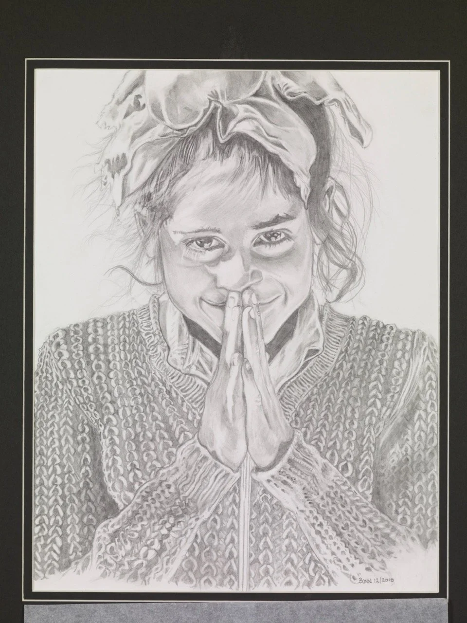A detailed pencil portrait of a young girl with short hair, wearing a large bow headband, and a textured sweater, holding her hands together near her face in a prayer gesture.