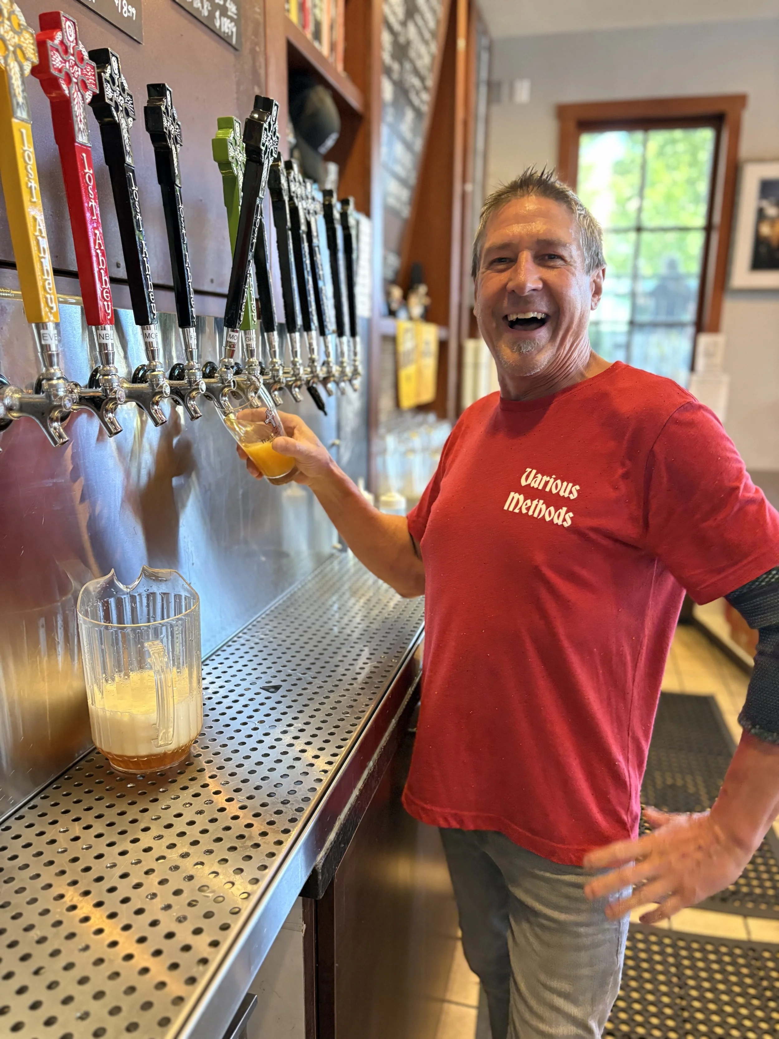 A man with short gray hair and a red shirt that says 'Various Methods,' pouring a beer from a tap at a bar or brewery, smiling and looking at the camera, with a background of several beer taps and a window with greenery outside.