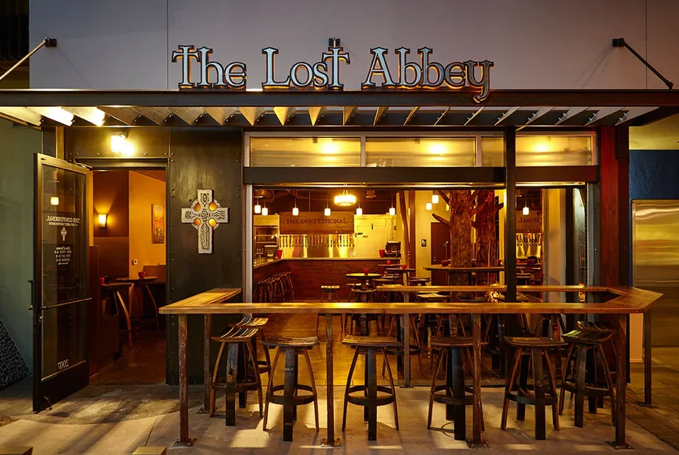 Exterior view of a restaurant named 'The Lost Abbey' with dark wood accents and a long standing table with stools outside, and a glimpse of the interior showing bar seating, warm lighting, and a decor theme inspired by craft beer.