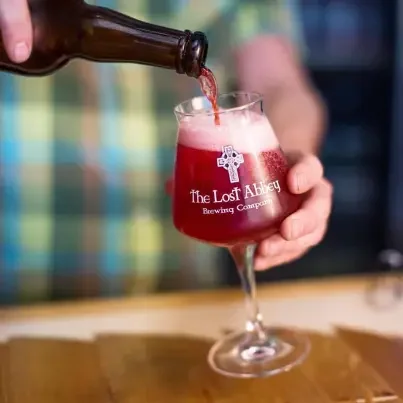 A person pouring a red beer with foam into a glass with the logo 'The Lost Abbey Brewing Company' on it.