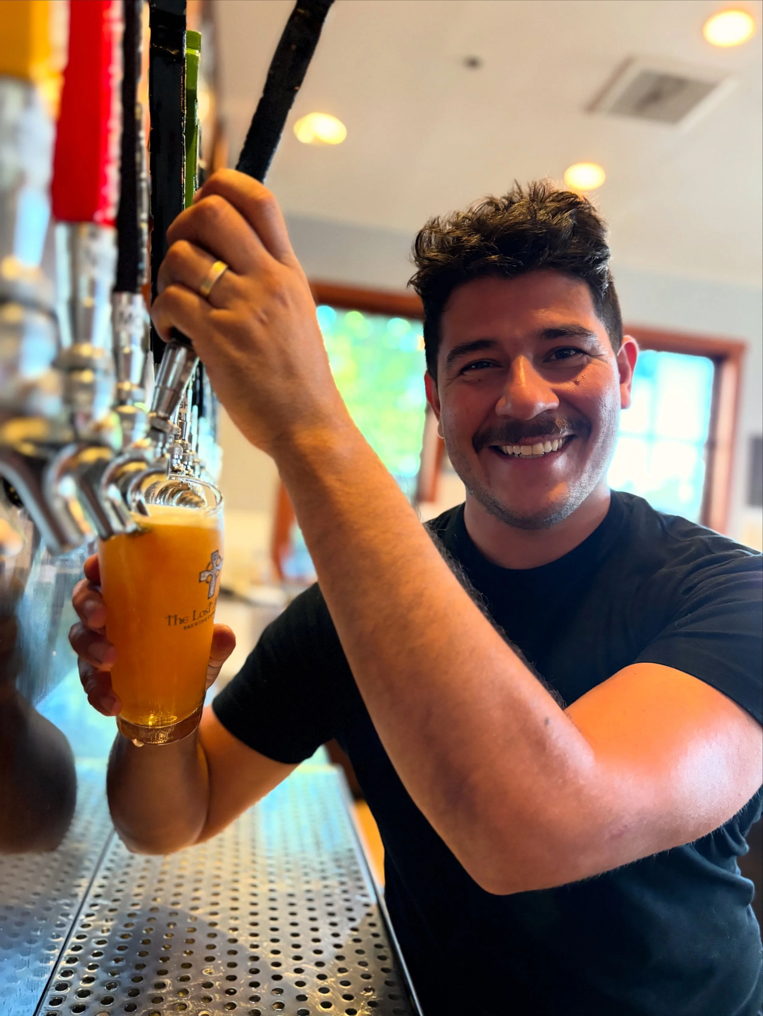 A smiling man in a black t-shirt with short dark hair pours beer from a tap at a bar. He holds a glass with an orange logo, and there are several beer taps in front of him.