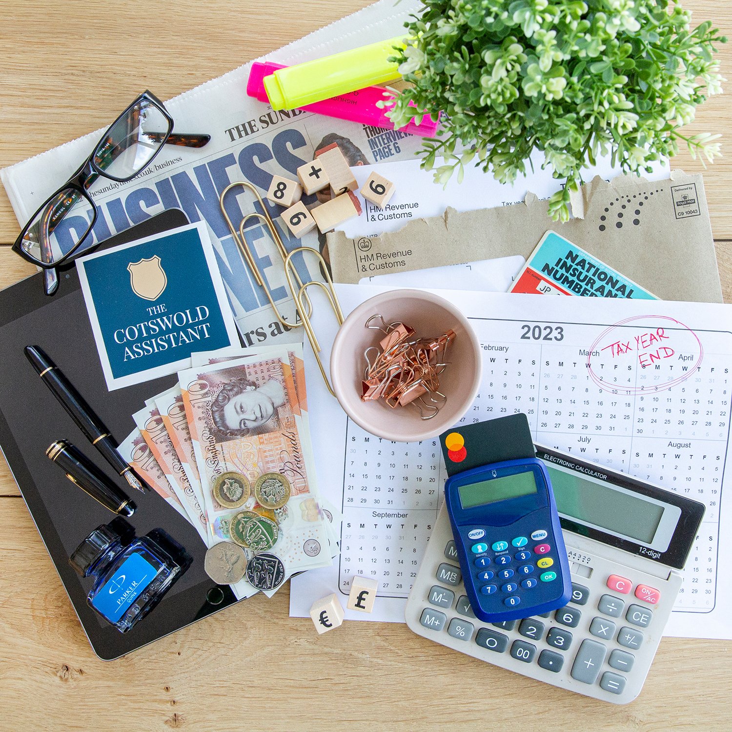 A cluttered desk with financial documents, cash, coins, a calculator, a pen, reading glasses, a small plant, a calendar marked with 'Tax Year End' on March, highlighters, paper clips, and a newspaper.