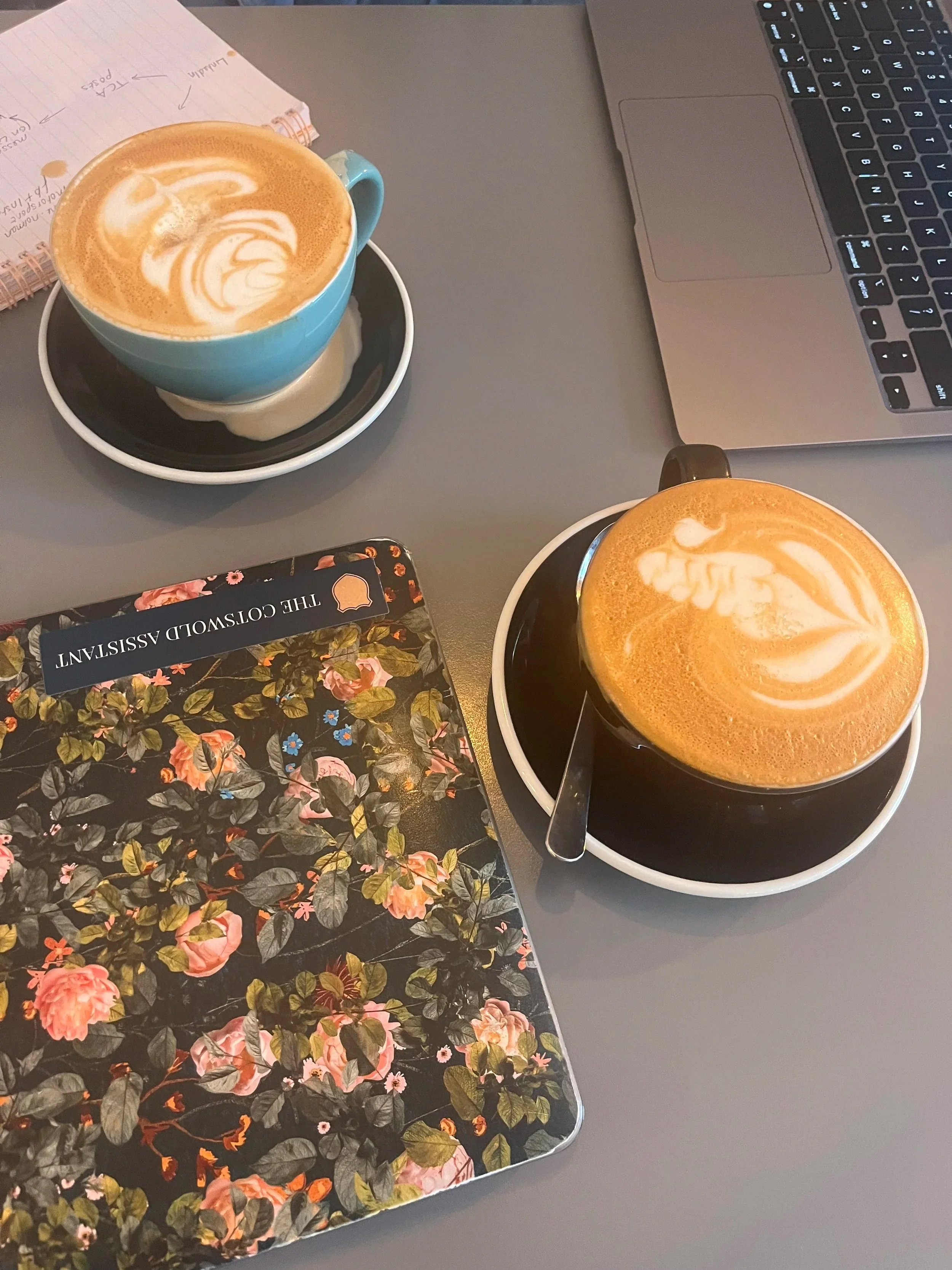 Two cups of coffee with latte art on top, placed on a gray table beside a laptop, a floral planner, and a notebook.