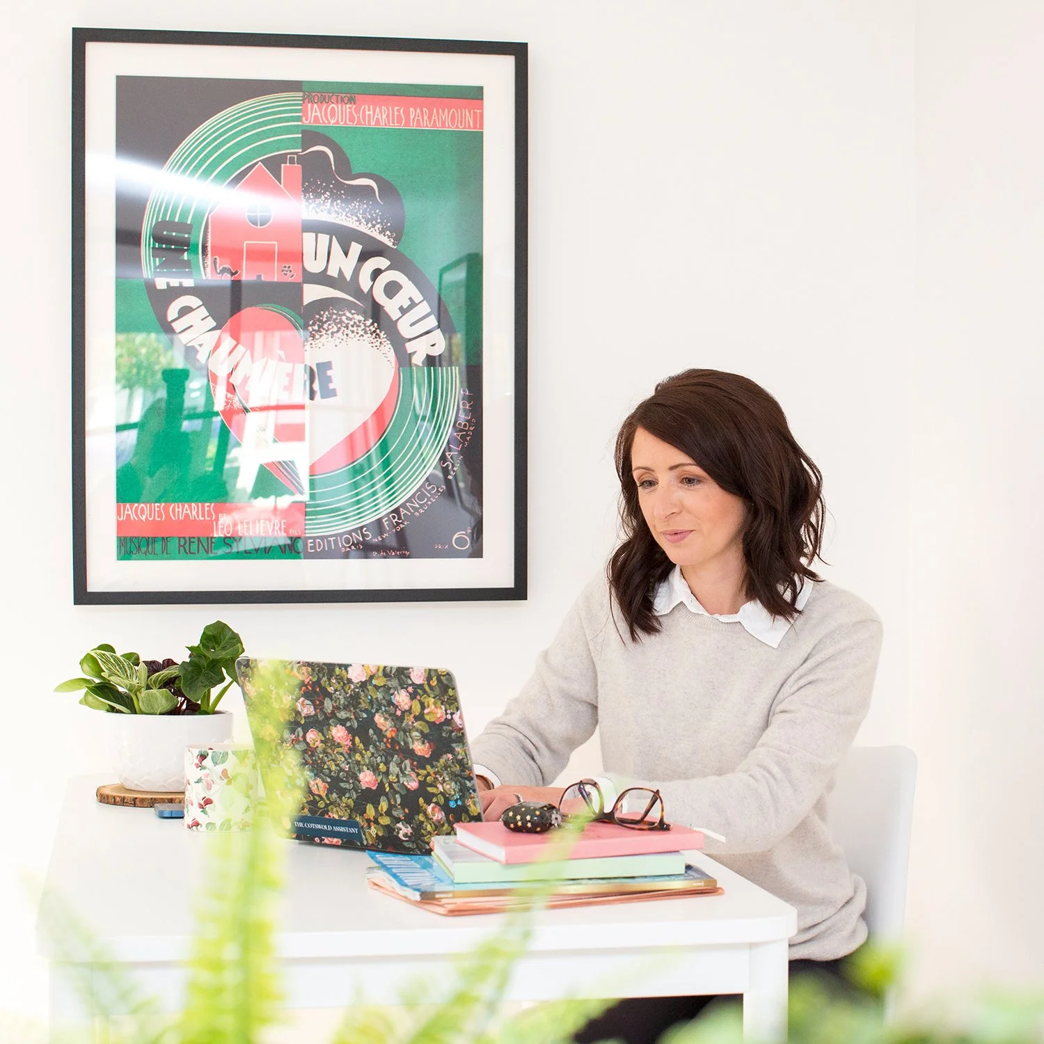 A woman with dark brown hair working on a laptop at a white desk in a bright room. There are colorful notebooks, a pair of glasses, and a small black object on the desk. A potted plant is on the left side of the desk. A poster with abstract art and text is hung on the white wall behind her.