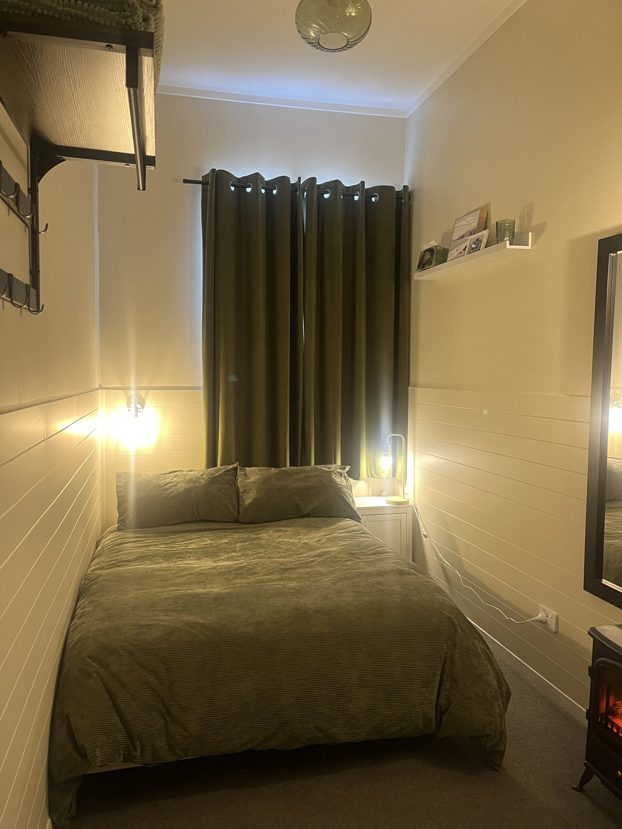 A cozy bedroom with a made bed covered in olive green bedding, illuminated by two bedside lamps. There are dark green curtains covering a window behind the bed, and a mirror on the right wall. A small shelf with picture frames and decor is visible above the bed, and a wall-mounted storage unit is on the left side of the image. A ceiling light fixture is also visible.