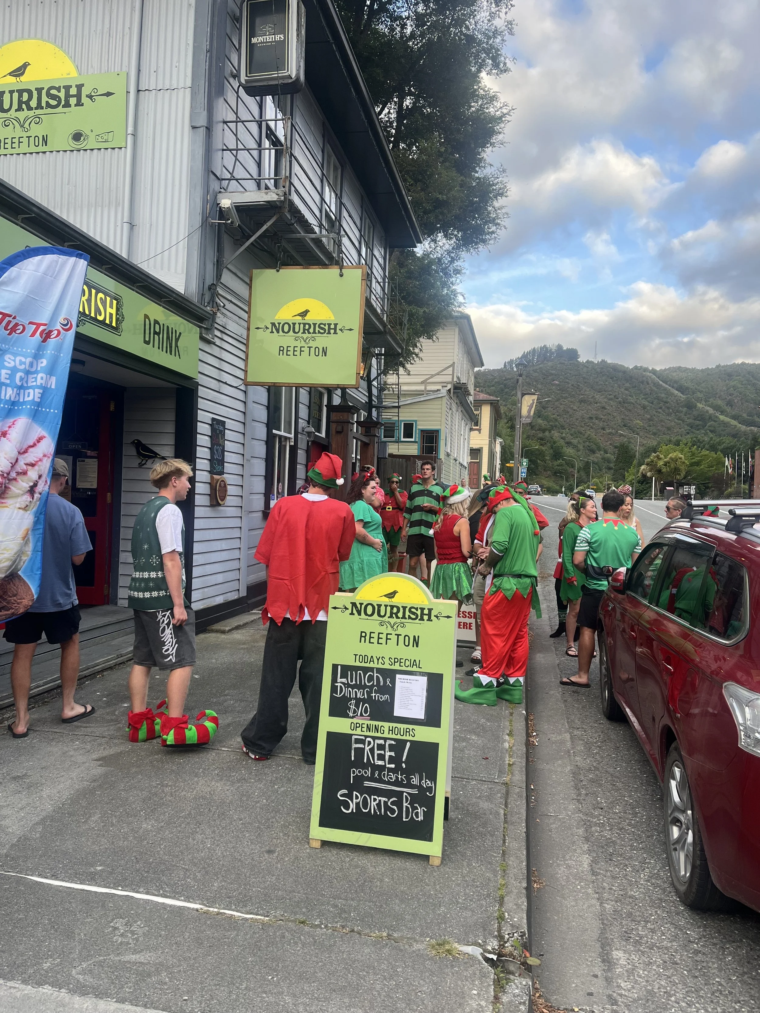 People dressed in Christmas-themed clothing and accessories outside a restaurant, with signs advertising food and drink specials, and a mountain landscape in the background.