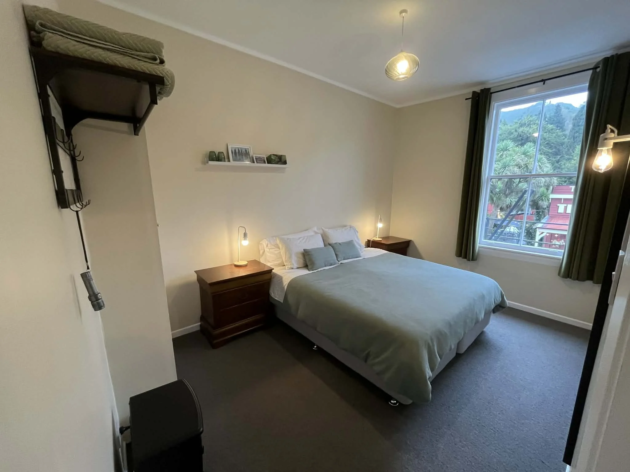 A bedroom with a bed, a nightstand with a lamp, large window with green curtains, a shelf with picture frames, and a wall-mounted coat rack with folded blankets.