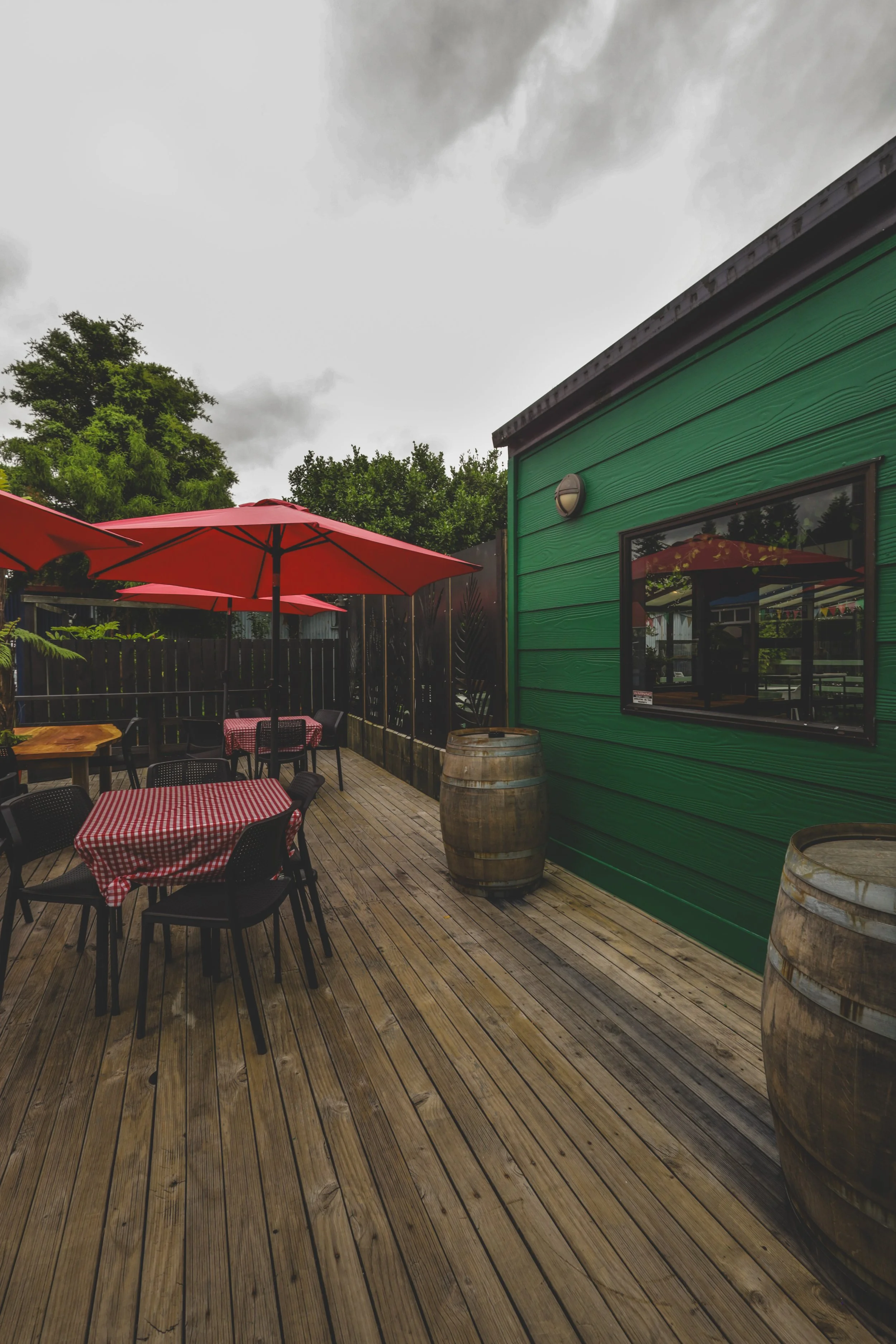 Outdoor patio area with tables covered in red checkered tablecloths, red umbrellas, wooden barrels, and a green building with a window on a cloudy day.