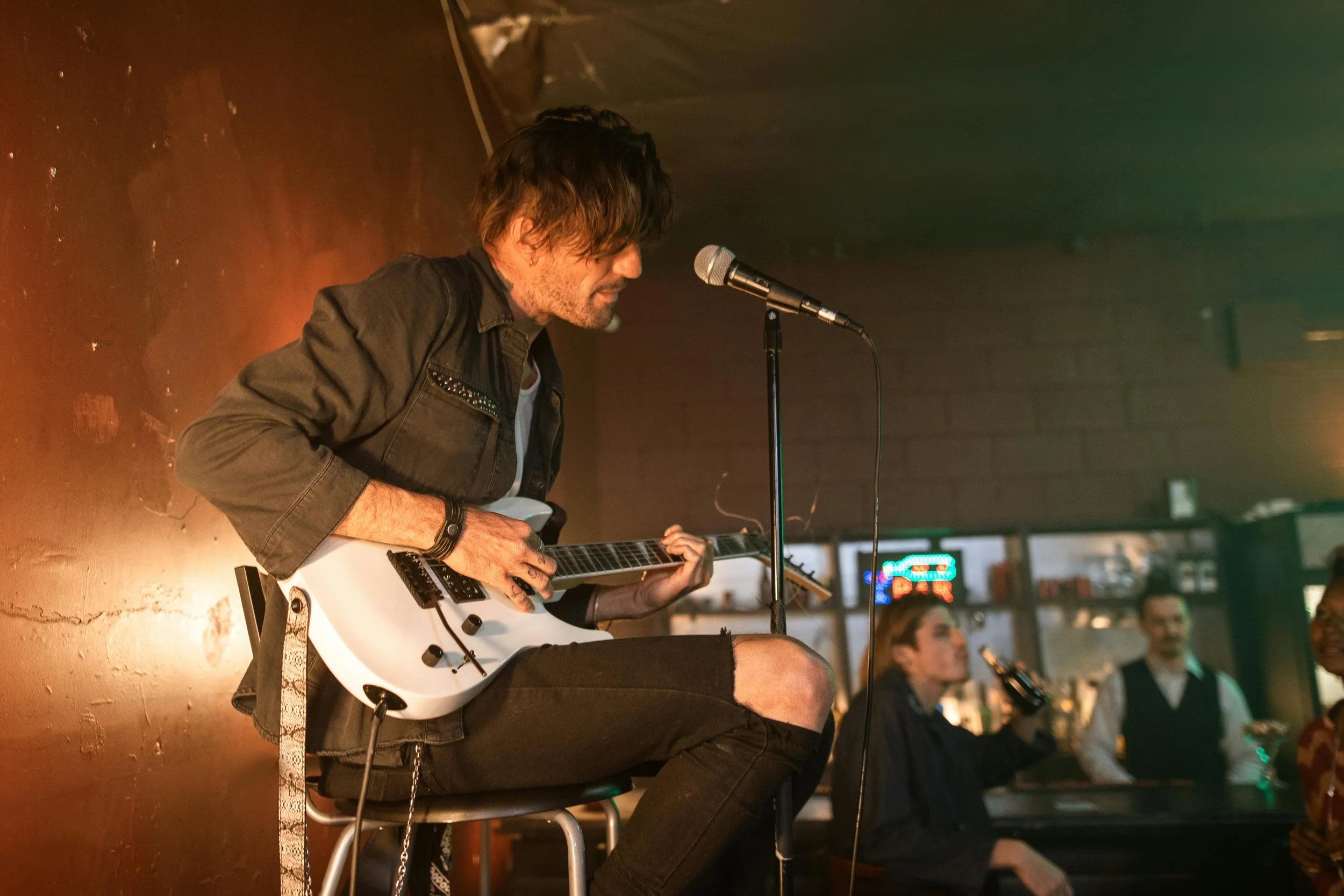 A male musician with shaggy hair, wearing a black jacket, is sitting on a stool playing an electric guitar in a dimly lit bar or music venue, with a woman and another man in the background.