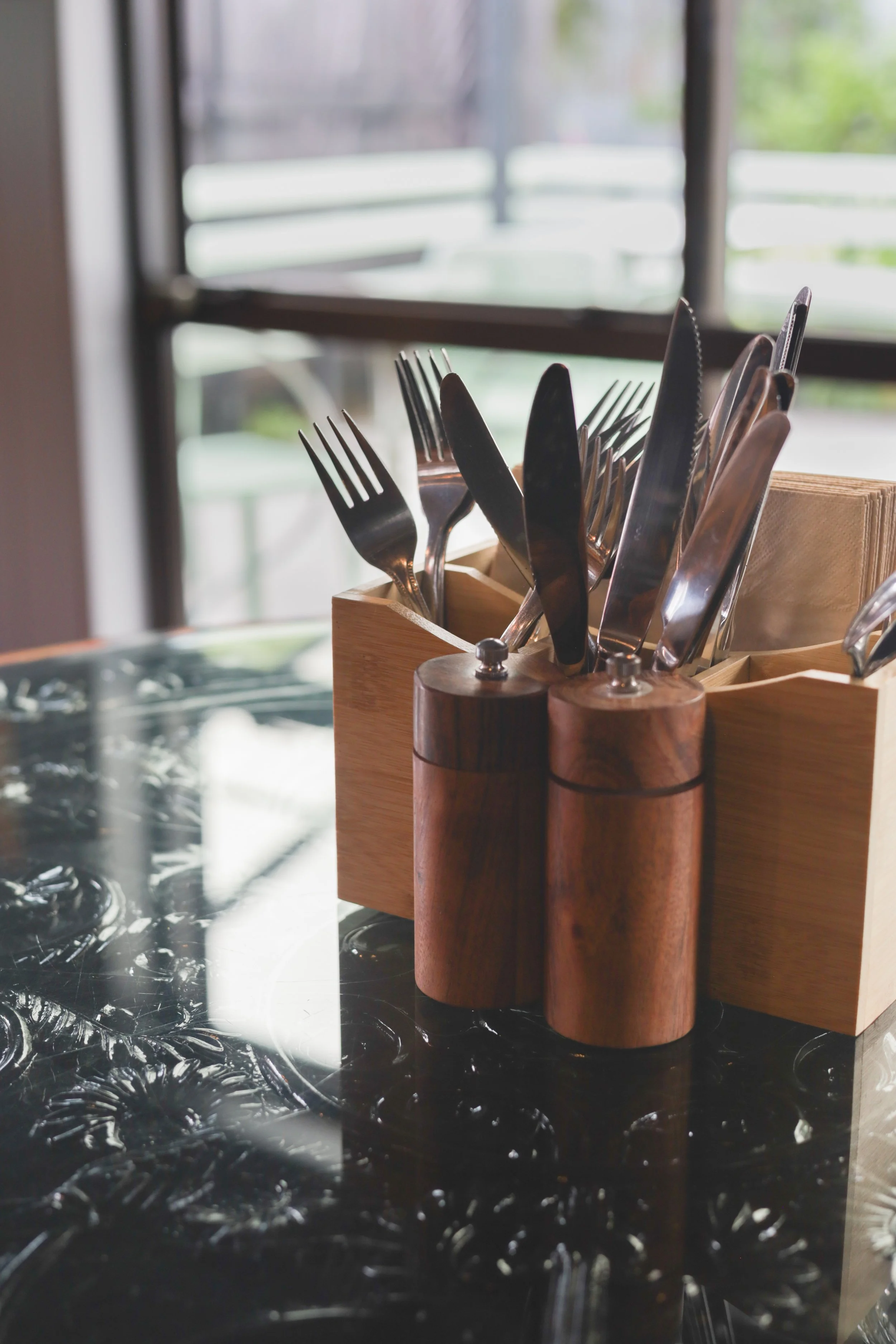 Cutlery set including forks, knives, and spoons in a wooden caddy on a black decorative table near a window.