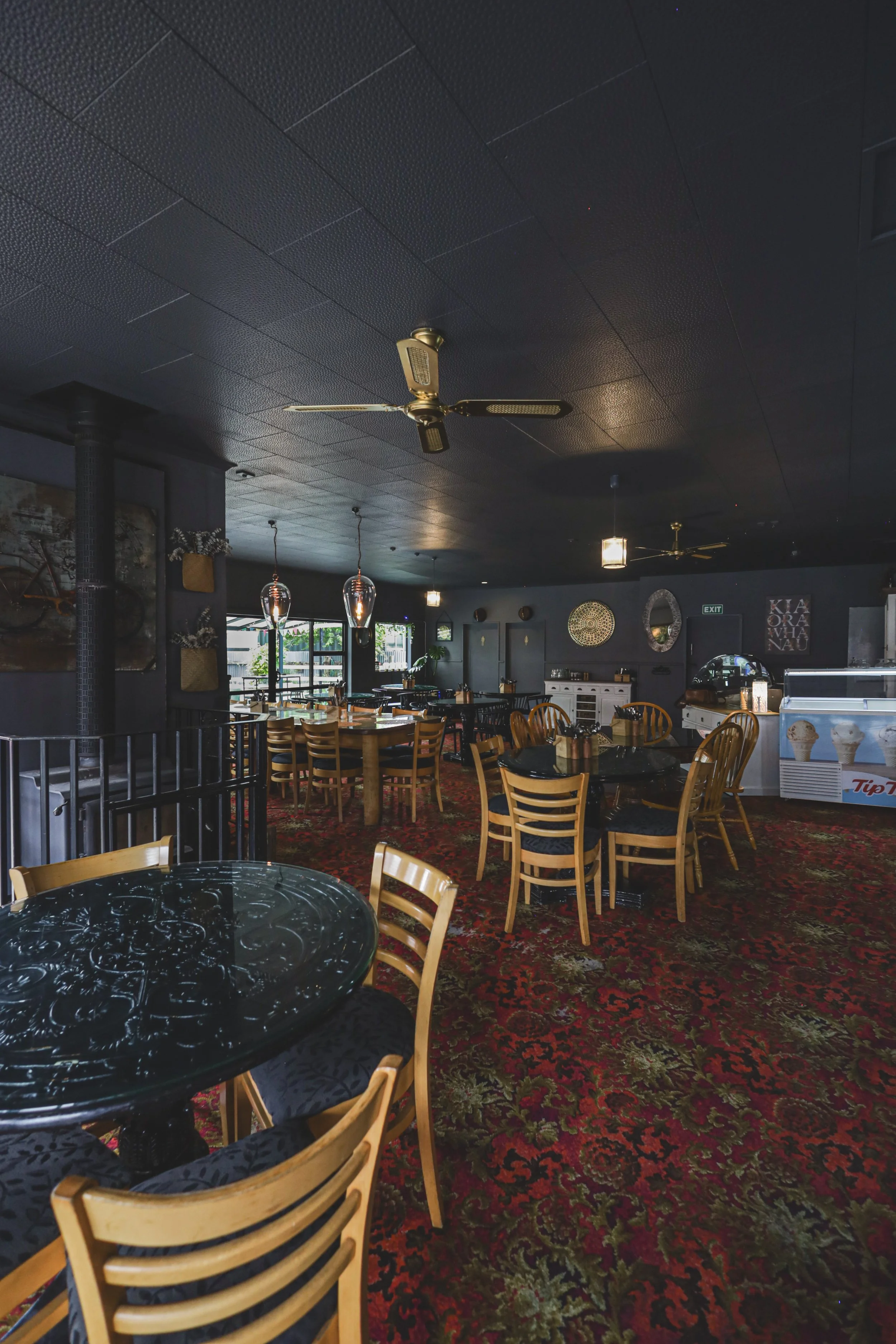 Interior of a dimly lit restaurant with wooden chairs, black tables, a red and green floral carpet, pendant lights, ceiling fans, and an ice cream freezer.
