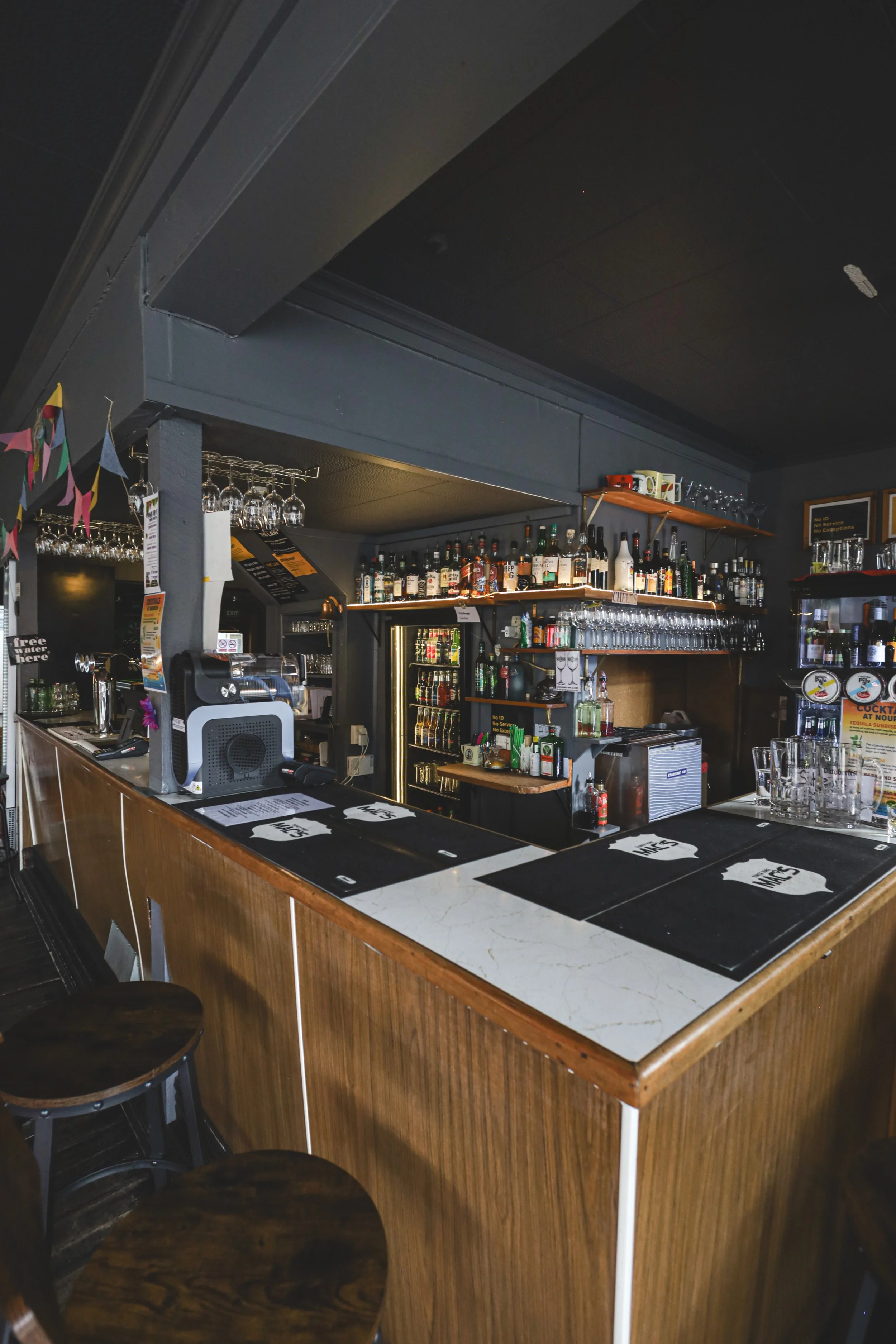 Empty bar counter with bar mats, glasses, and shelves stocked with bottles of alcohol in a dimly lit bar or pub.