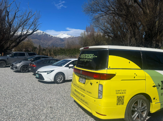 Parking lot with multiple cars, yellow bus, trees, mountain range, and blue sky in the background.