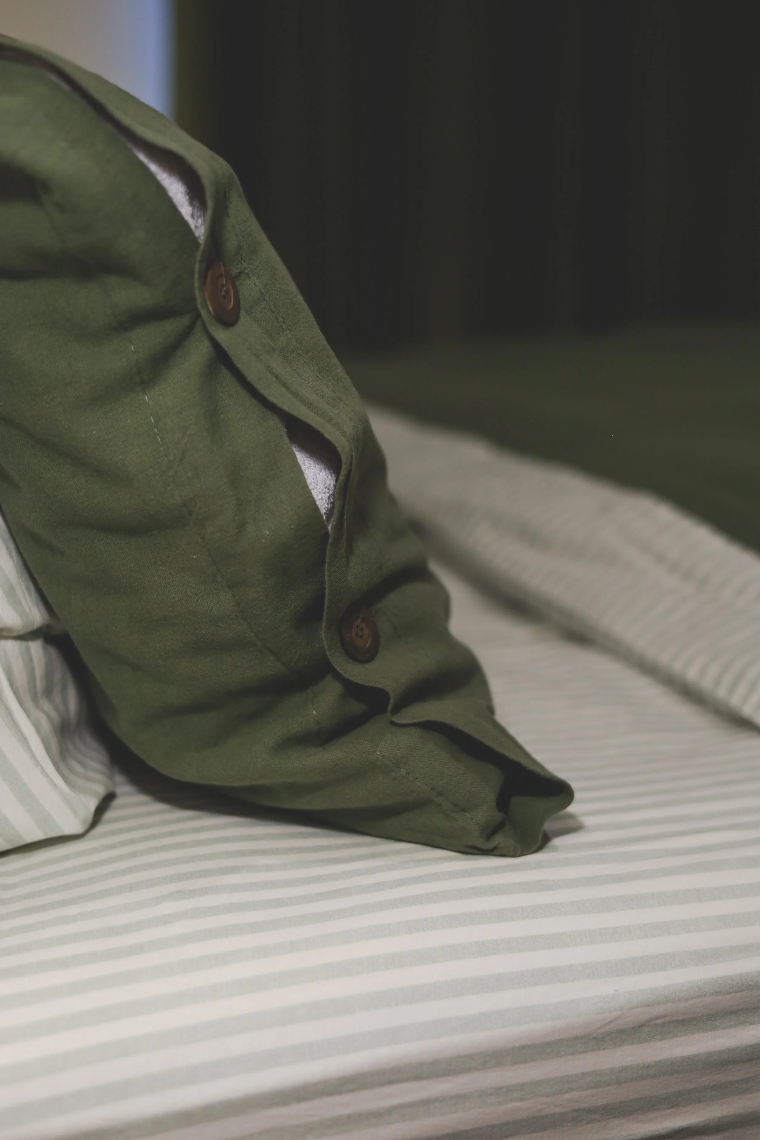 Close-up of a green button-up shirt with brown buttons, lying on a bed with a striped sheet.