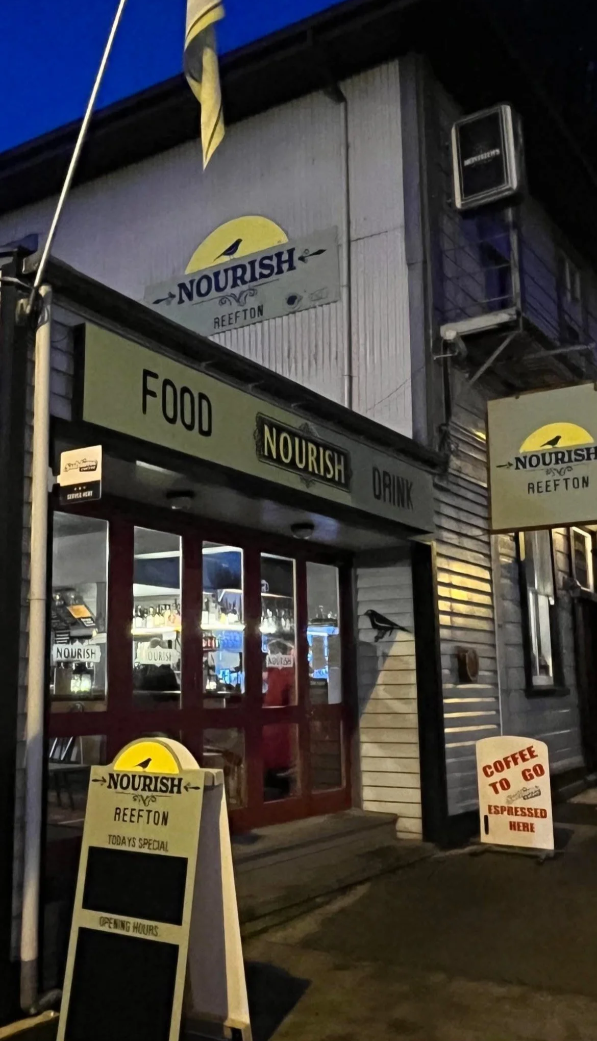 Nighttime exterior of a restaurant named Nourish Reefton, featuring signs promoting food, drinks, and coffee, with a chalkboard menu and promotional sandwich board outside.