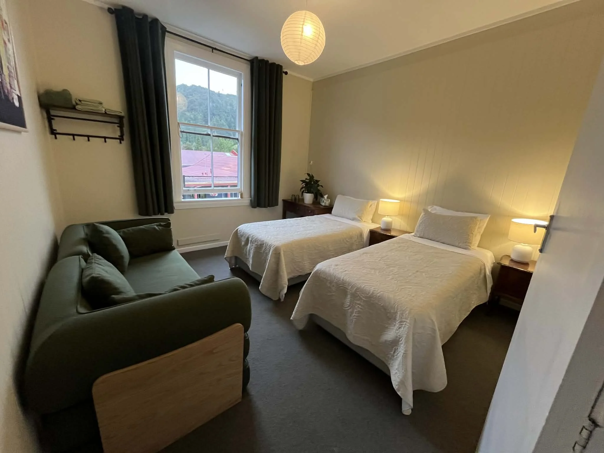 A cozy bedroom with two twin beds covered in white textured bedspreads, nightstands with lamps, a green armchair, a window with dark curtains, a shelf with towels, and a ceiling light with a paper lantern design.