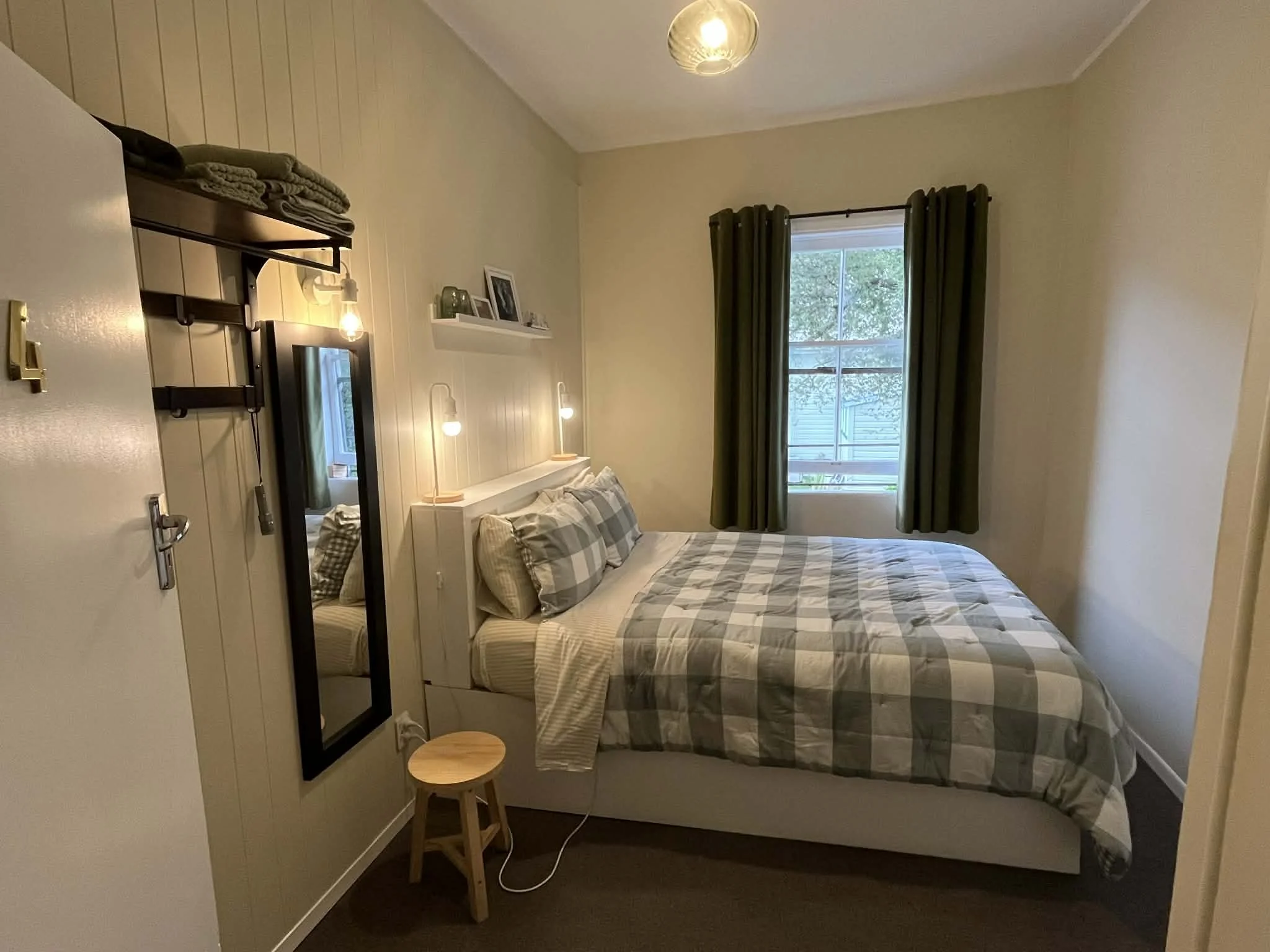 A cozy bedroom with beige walls, a bed with checkered bedding, a window with dark green curtains, a small wooden stool, and decorative items on a white shelf.