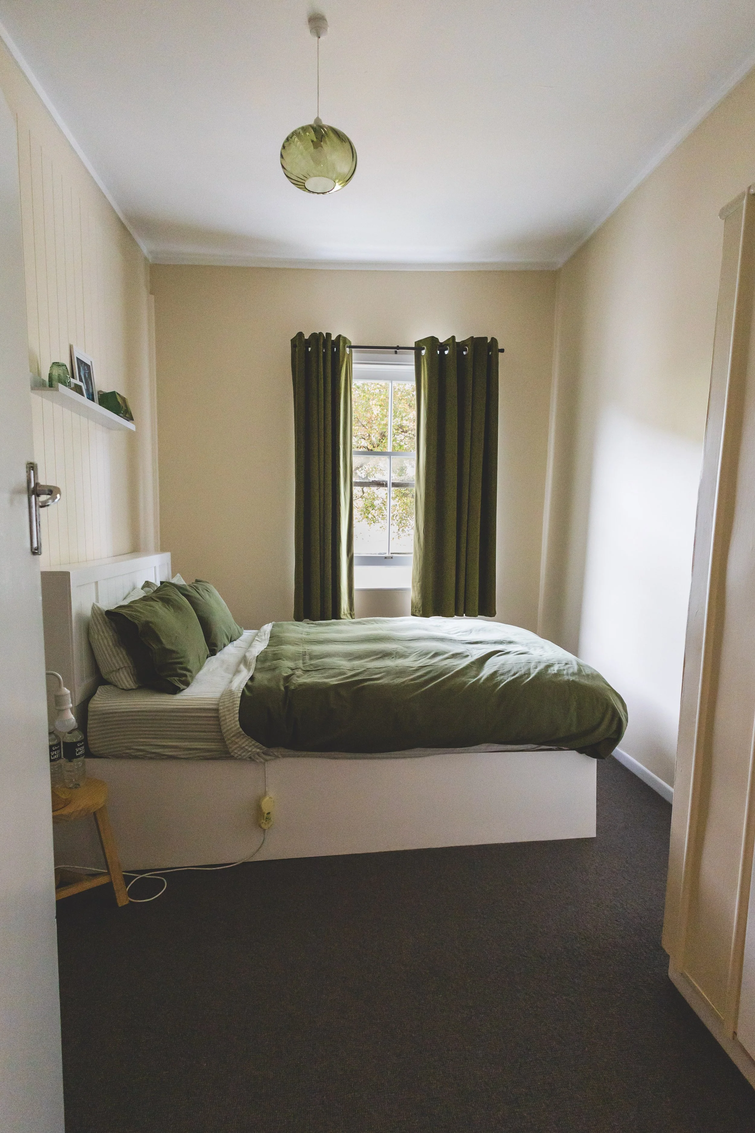 A bedroom with a bed covered in green bedding, a window with green curtains, a small bedside table with bottles, and a ceiling light