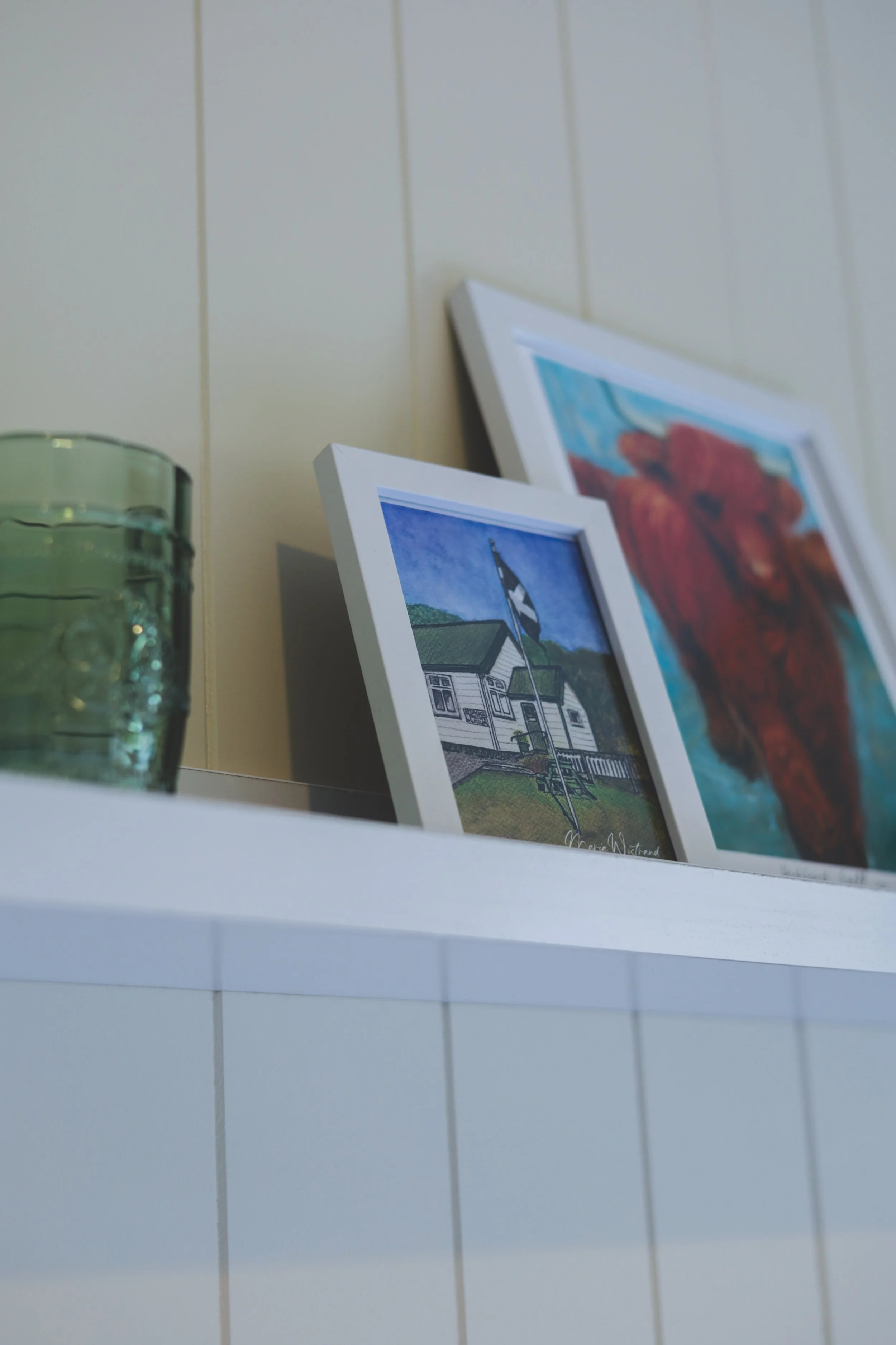 Shelf with two framed paintings and a green glass candle holder.