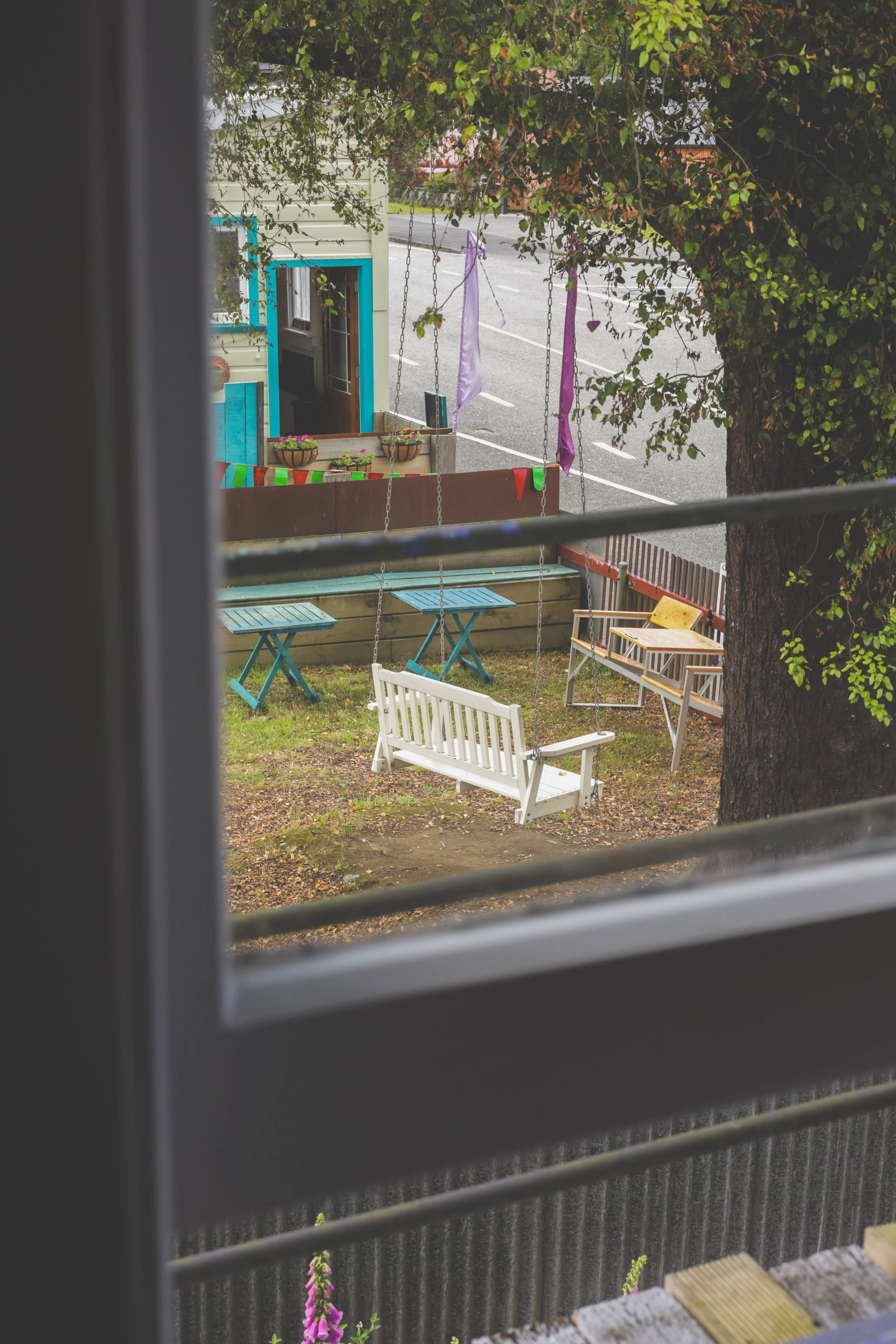 View of a backyard area seen through a window with horizontal blinds, featuring a white swing, a wooden bench, small tables, potted plants, and colorful ribbons hanging from a tree branch.