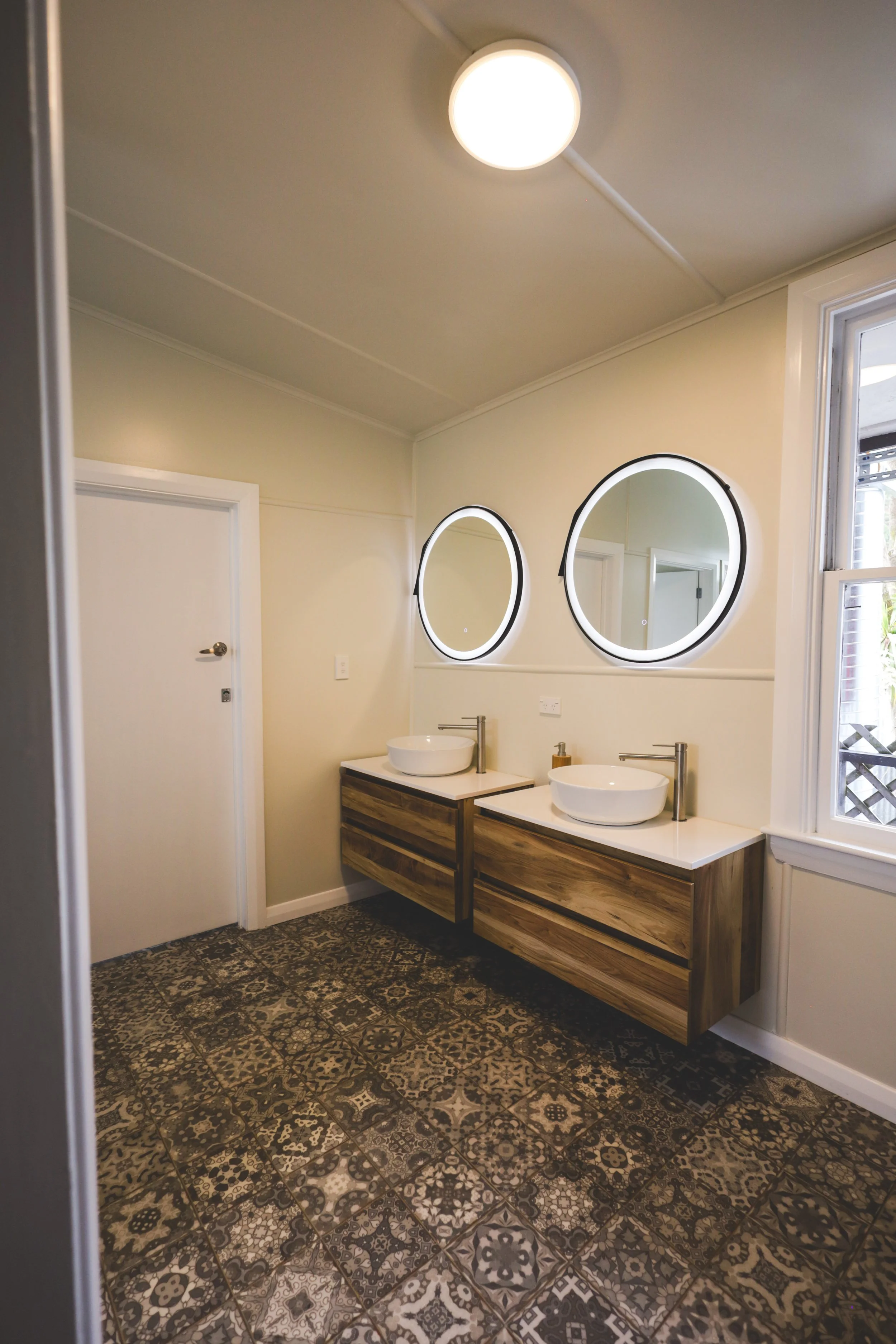 Bathroom with two round illuminated mirrors above two wooden vanities, each with a modern vessel sink, a soap dispenser, and a large window to the right, with patterned tile flooring and a ceiling light.