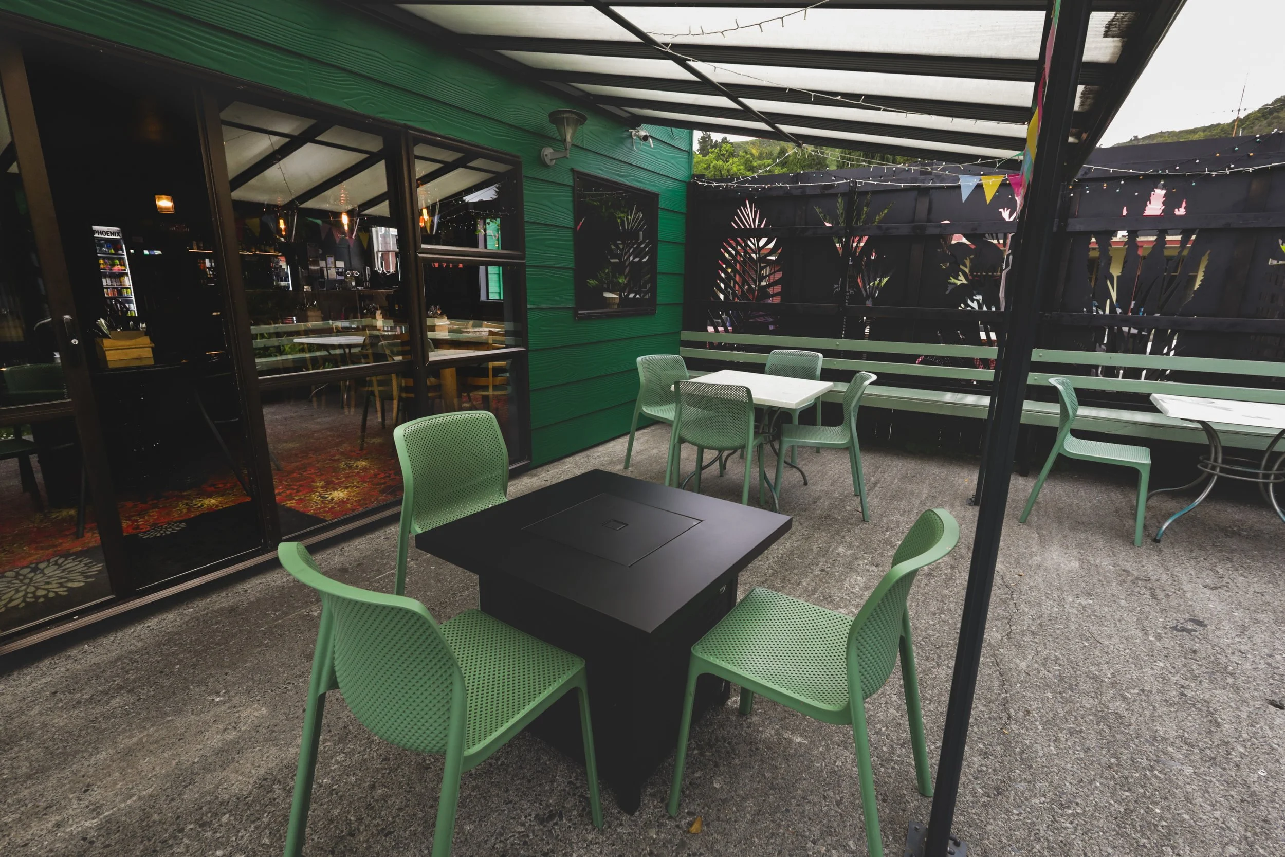 Outdoor patio with four green chairs around a black fire pit table and other white tables and chairs, enclosed by a black fence with colorful bunting, adjacent to a building with green siding and large glass windows.