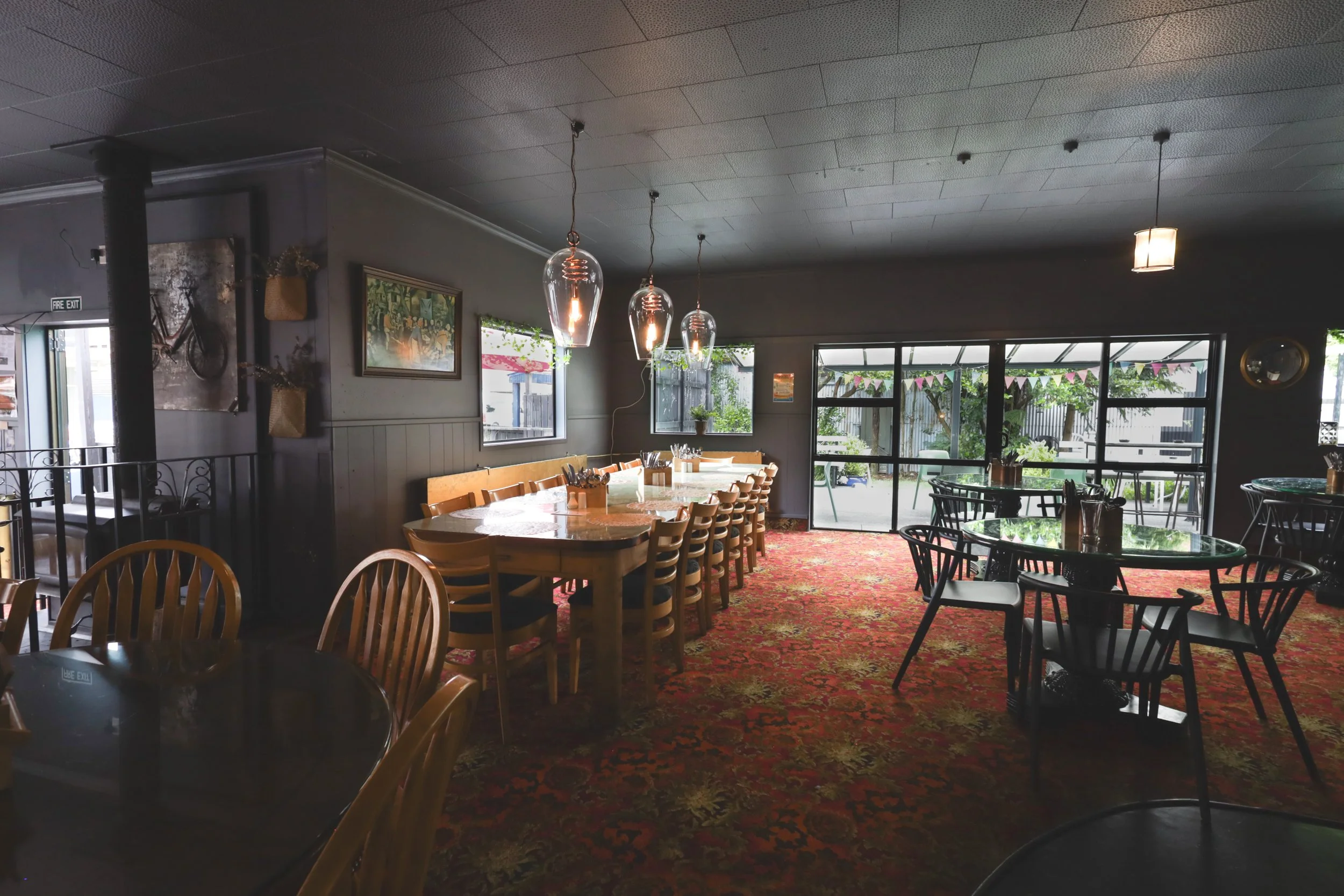 Interior view of a cozy cafe with wooden and black chairs around tables, pendant lights hanging from a dark ceiling, large windows letting in natural light, and a richly patterned red carpet.