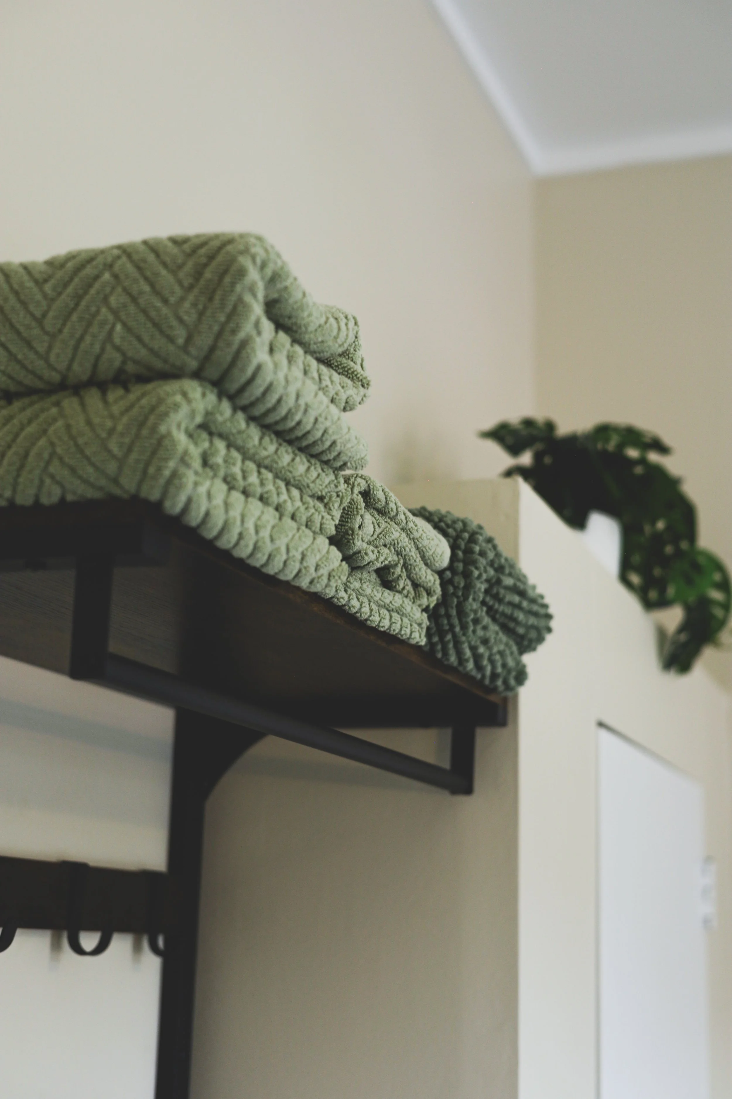 Stacked folded green patterned blankets on a black wall-mounted shelf with a small houseplant in a pot in the background.