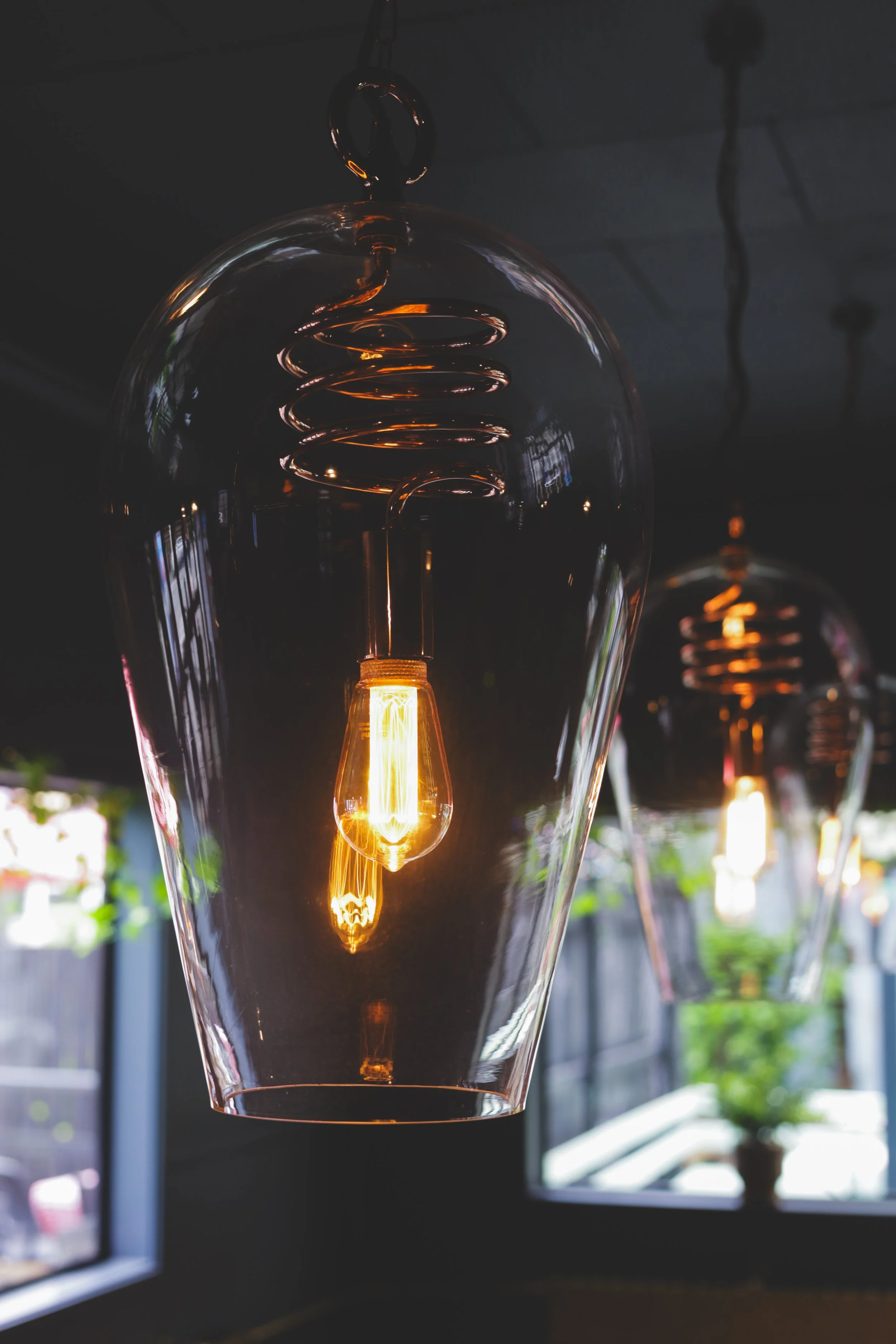 Close-up of hanging glass pendant light with an exposed filament bulb, with another similar light in the background, in a dimly lit interior space.