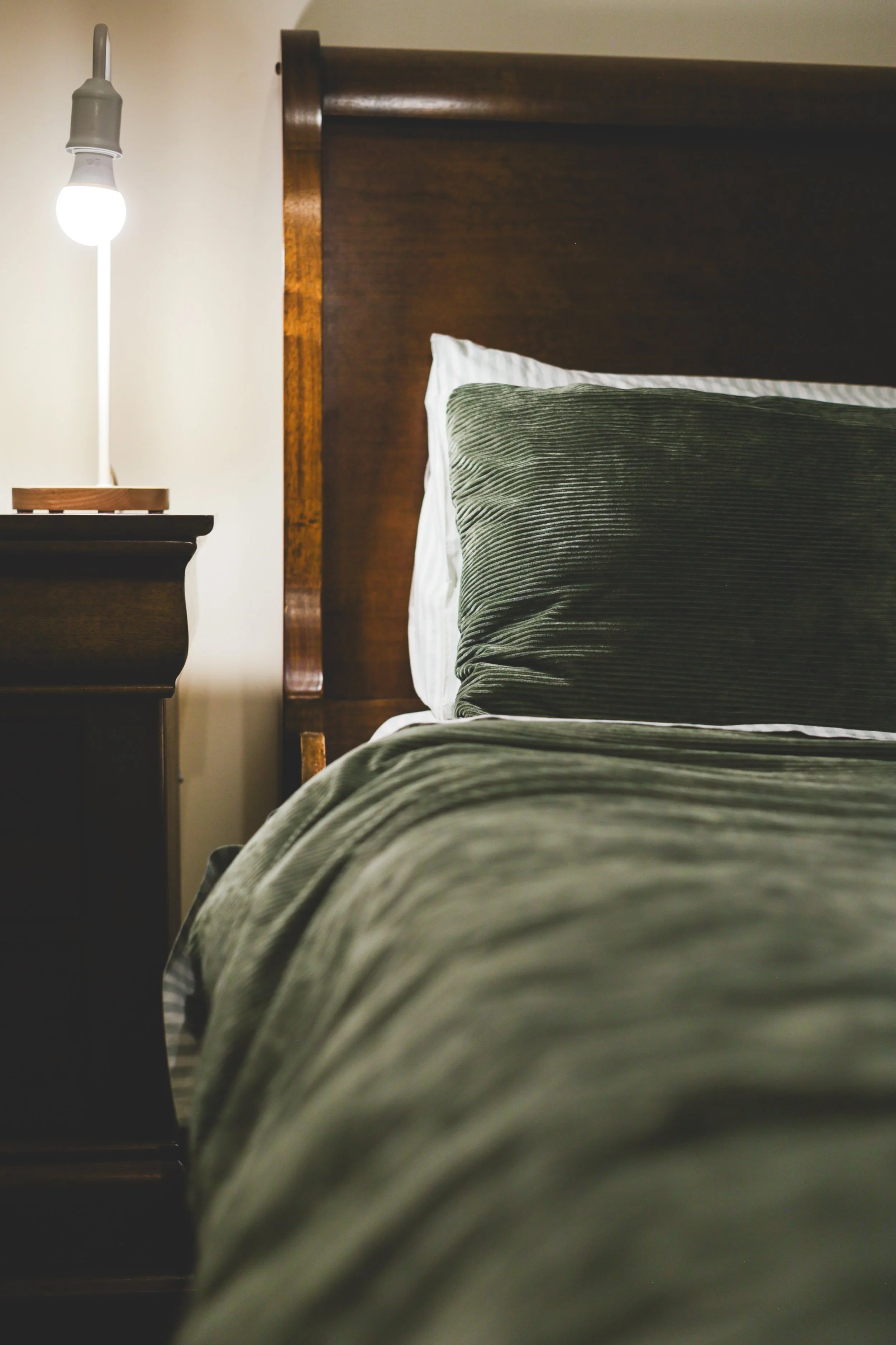 A neatly made bed with green striped sheets and pillow, a wooden headboard, and a dark bedside table with a modern lamp in a cozy bedroom.