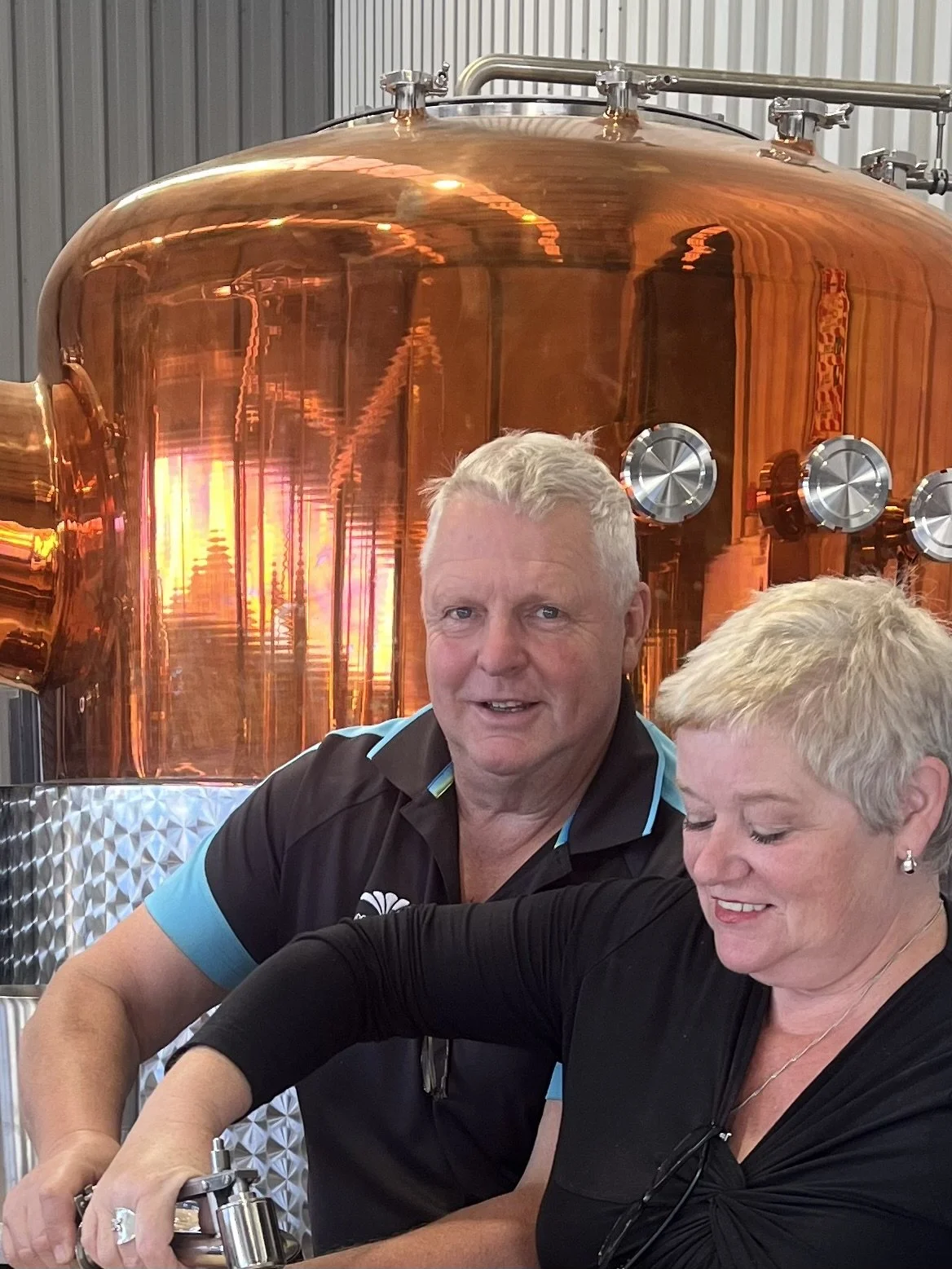 Two smiling people, a man and a woman, in front of a large copper distillation tank.