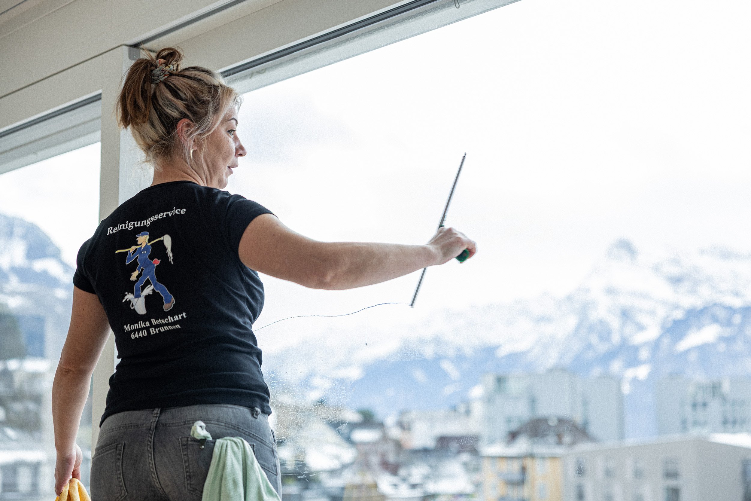Frau putzt eine große Glasfront mit einem Rakel, Blick auf schneebedeckte Berge im Hintergrund.