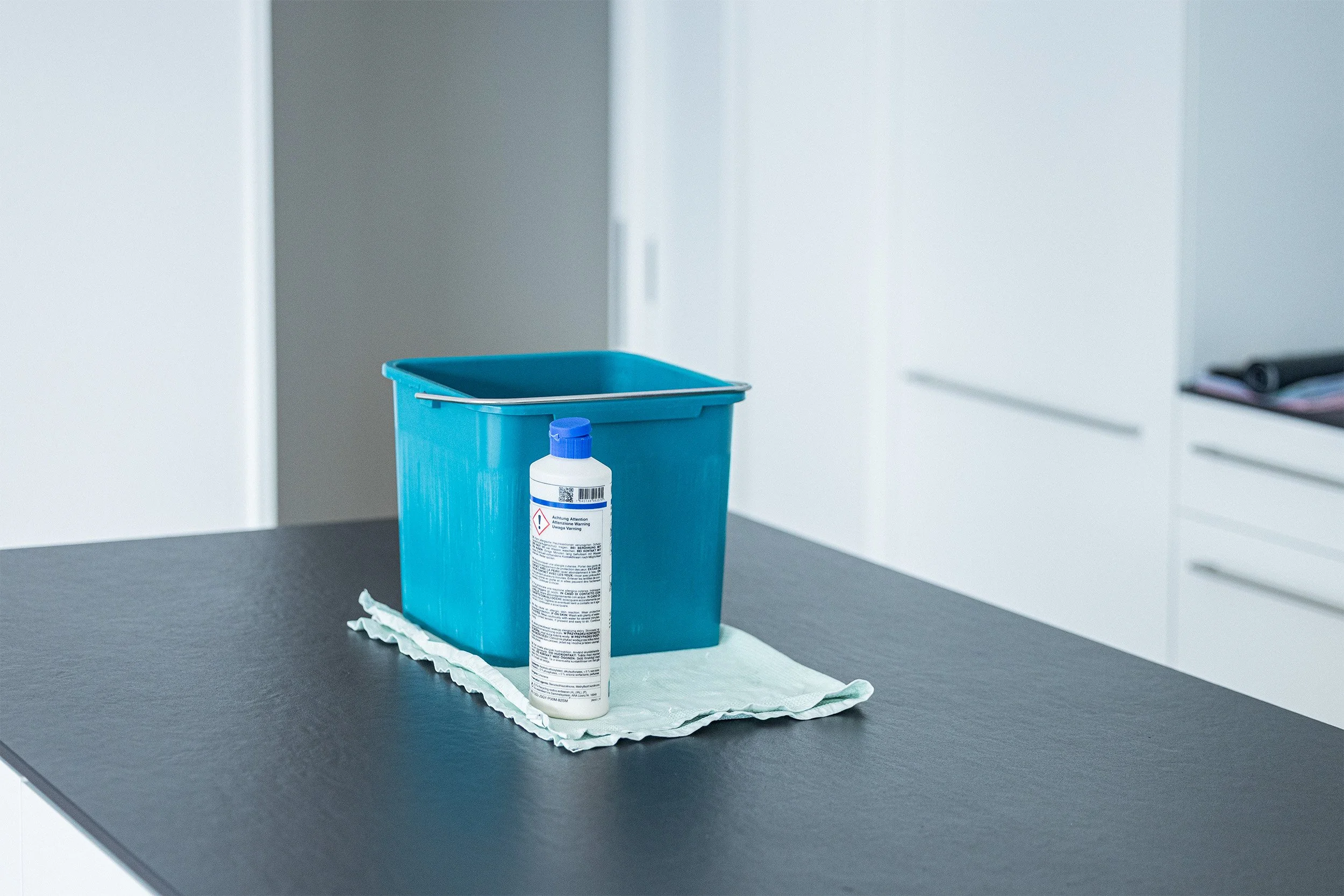 Ein blauer Mülleimer mit einer weißen Flasche Desinfektionsmittel auf einem weißen Tuch auf einem dunklen Tisch in einer modernen weißen Küche.