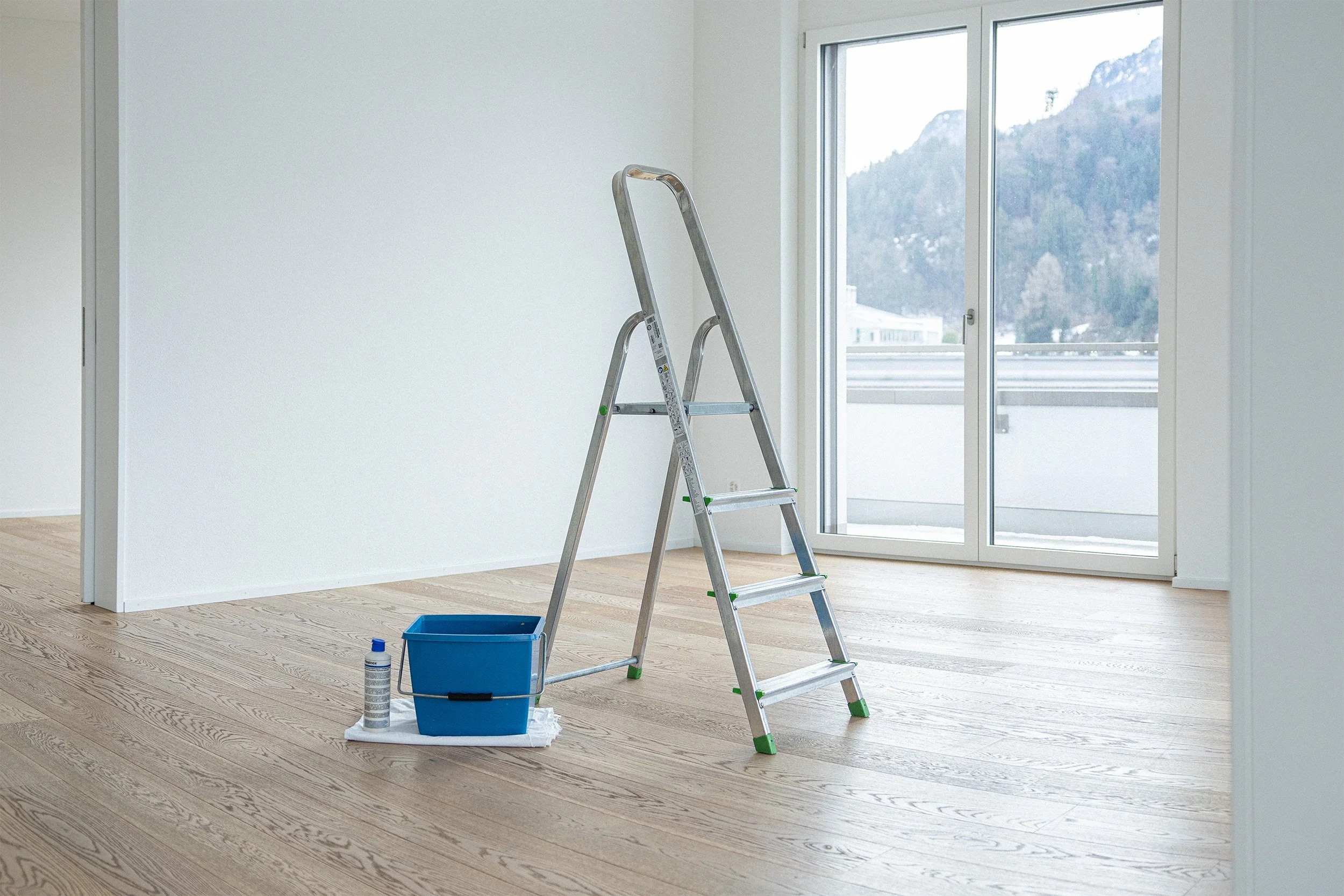 Un leeres Zimmer mit Holzboden, eine Leiter, eine blau Kunststoffbox, einen Lappen und eine Reinigungsflasche auf dem Boden, große Fenster mit Blick auf Berge im Hintergrund.