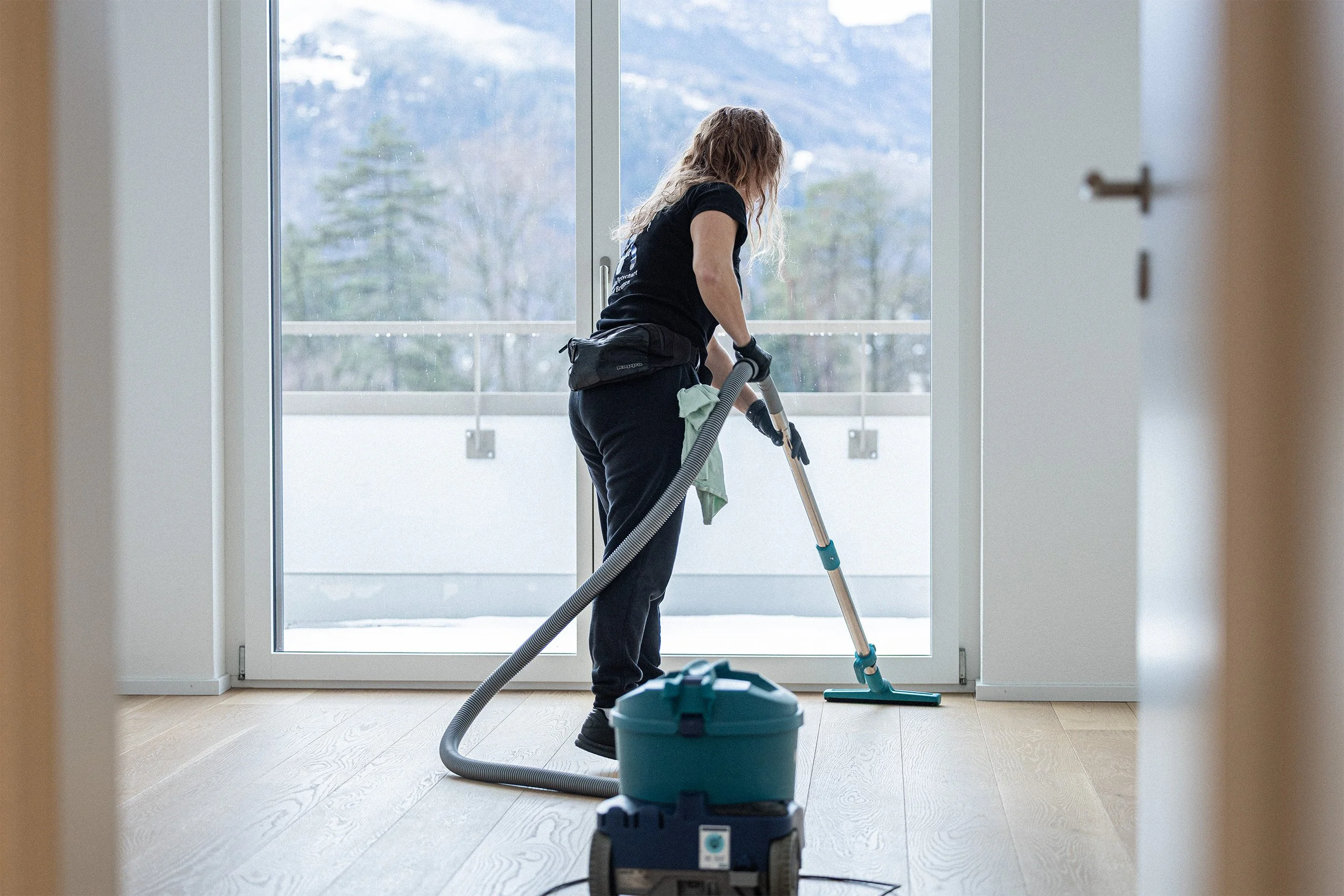 Eine Frau reinigt mit einem Staubsauger einen hellen Holzboden vor einer großen Glastür auf einem Balkon, mit einer Winterlandschaft im Hintergrund.