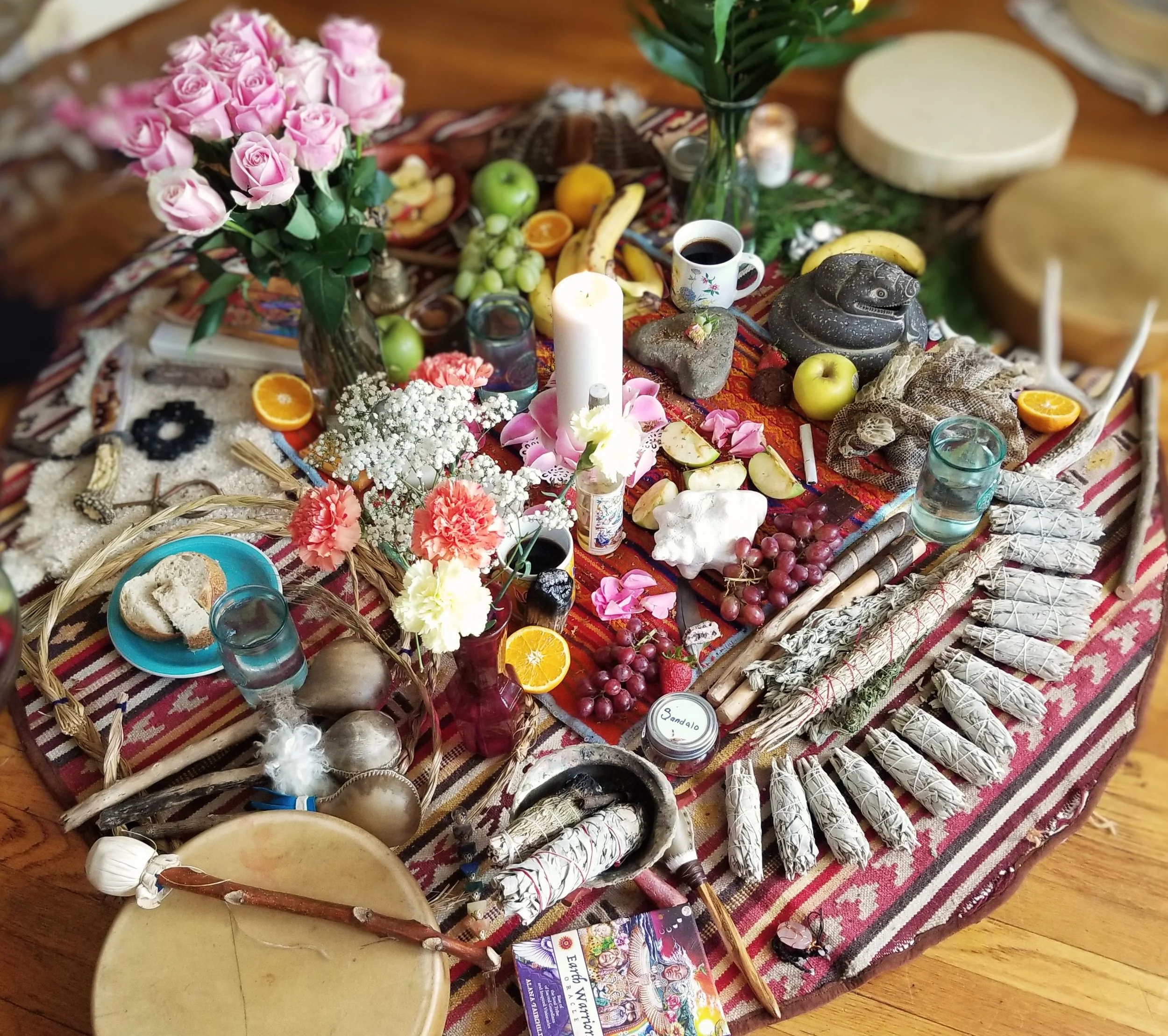 A colorful, eclectic table setting featuring flowers, fruits, candles, and decorative objects, with rolled papers and various artistic items on a patterned cloth.