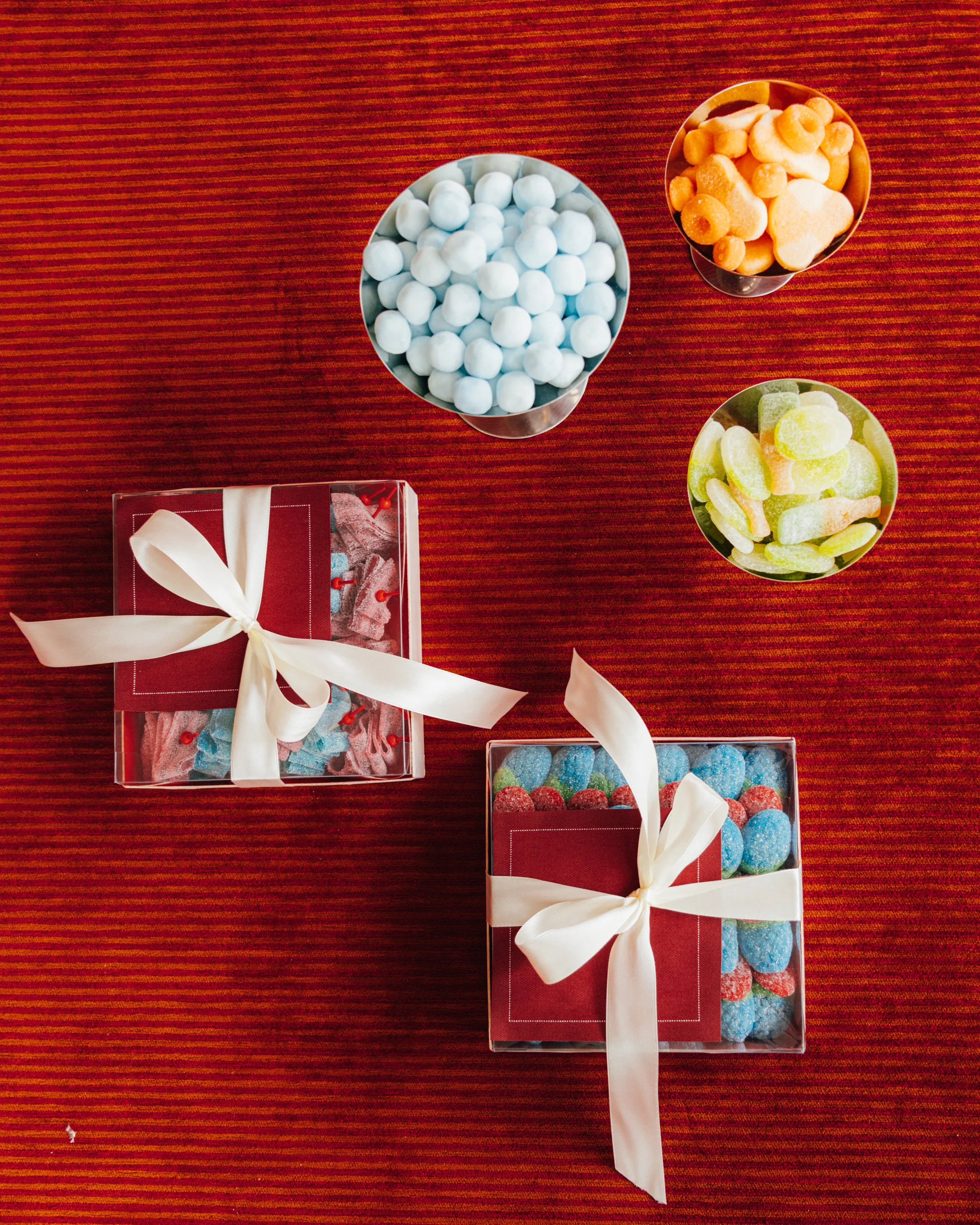 Three gift boxes with candies on a red striped tablecloth. The boxes are wrapped with white ribbons and each contains different colorful candies in small containers.