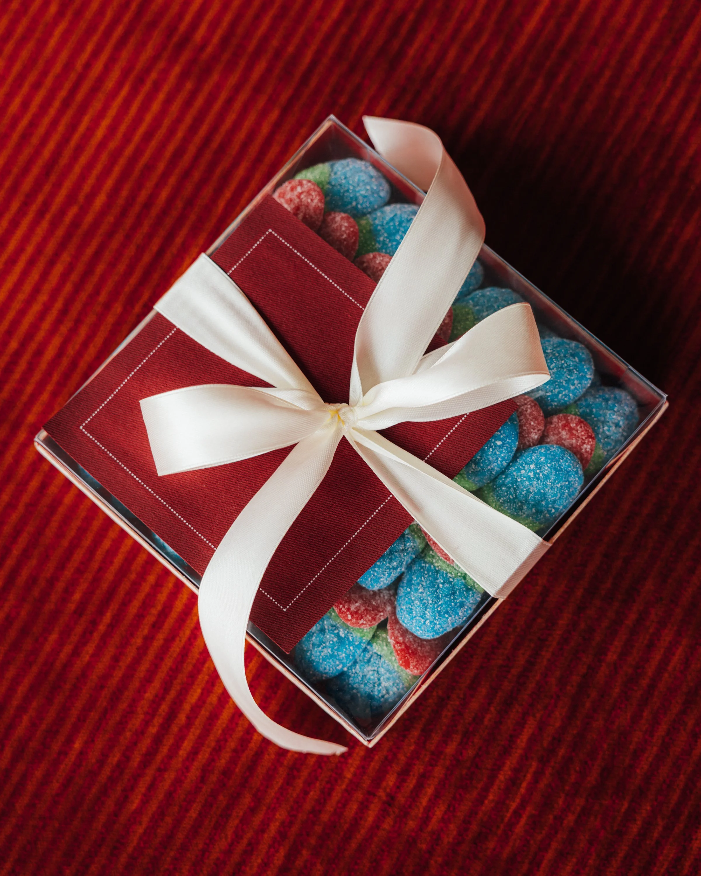 A red lacquered bento box filled with colorful sugar-coated candies, decorated with a large white satin bow, resting on a red-striped fabric surface. This is a bento box offered by CANDOR candy for one hour delivery in New York City.