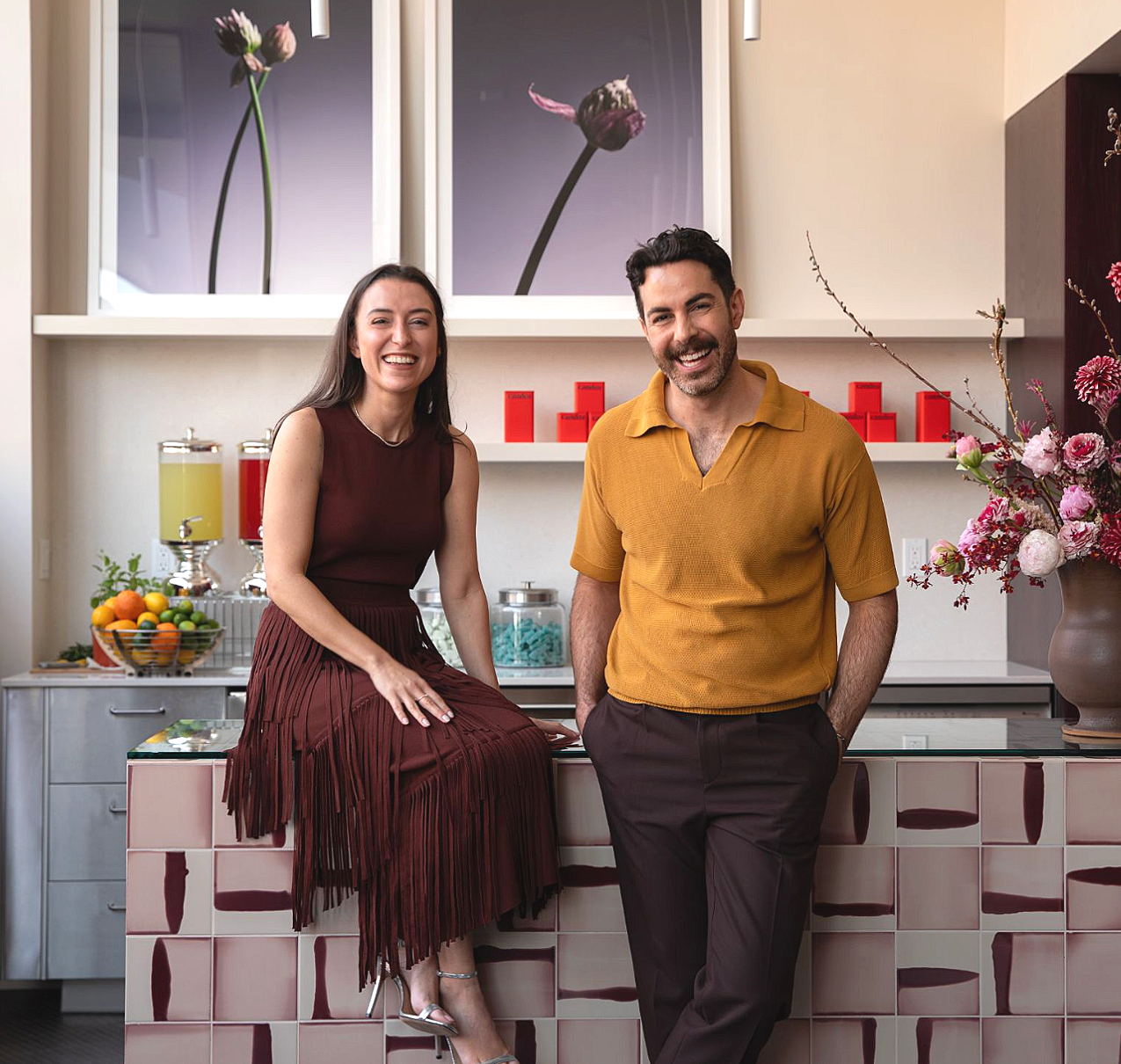 CANDOR founder Cat Cirino and CANDOR architect and designer Brian Cash are smiling near the distinct tiled checkout counter inside the shop - with floral and fruit decorations and framed floral artwork on the wall behind them.