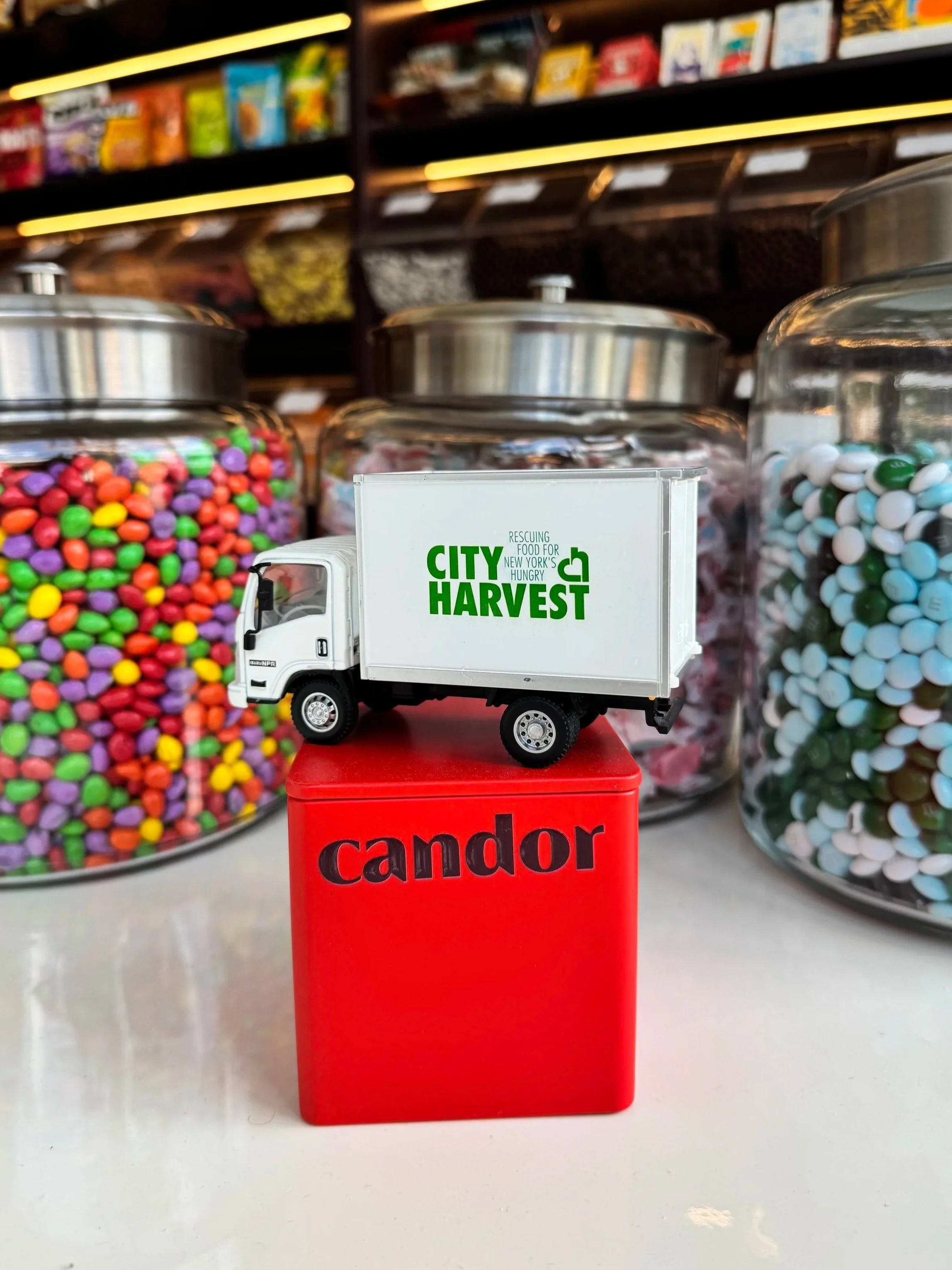 A small City Harvest toy truck sits atop a CANDOR tin, with CANDOR store shelves in the background - reflecting a partnership between the food pantry and the candy store.