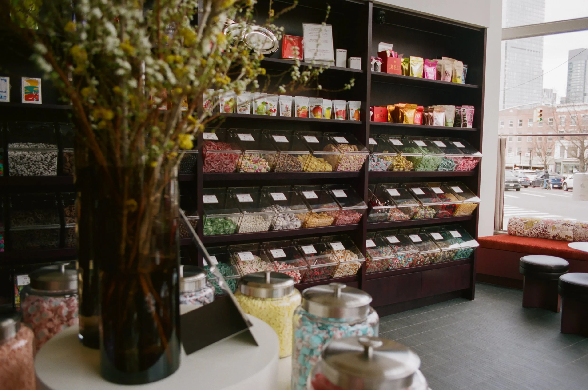 Indoor display of colorful candies and sweets in a store, with various packaged candies on shelves and jars of candies on a table in the foreground.