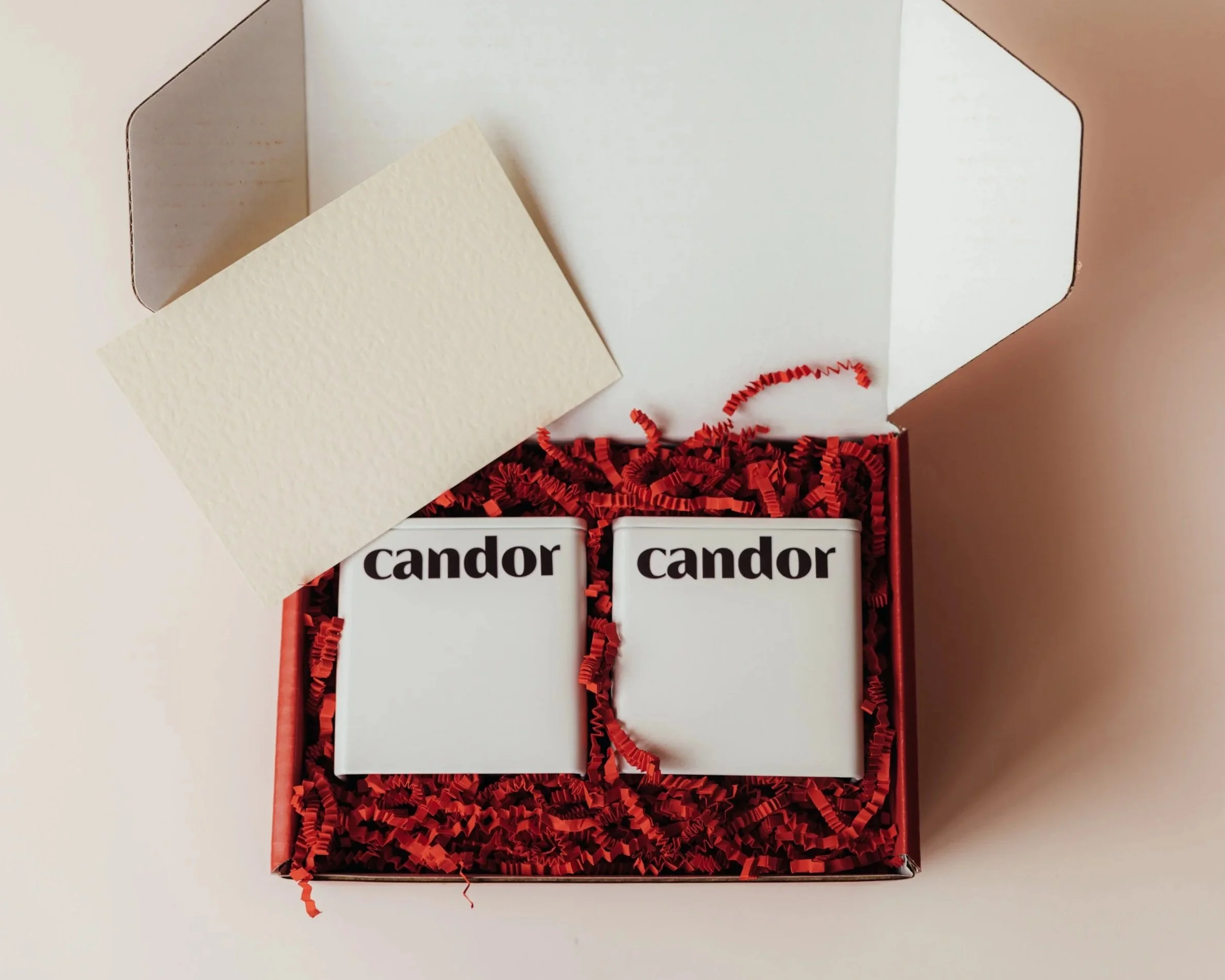 A red shipping box with shredded red paper filling, containing a two aluminum square tins with the word 'candor' printed on it in black letters. Shows a corporate gifting option offered by CANDOR - tins filled with candy or chocolate.