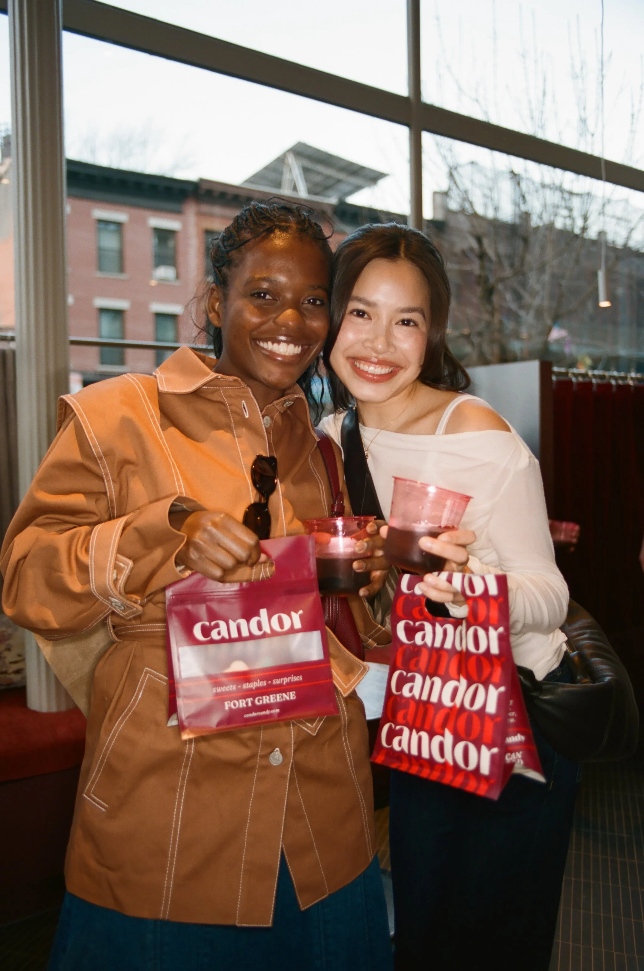 Two women smiling and holding drinks at an indoor event, with shopping bags from Candor and a red gift bag, in front of large windows showing trees and buildings outside.