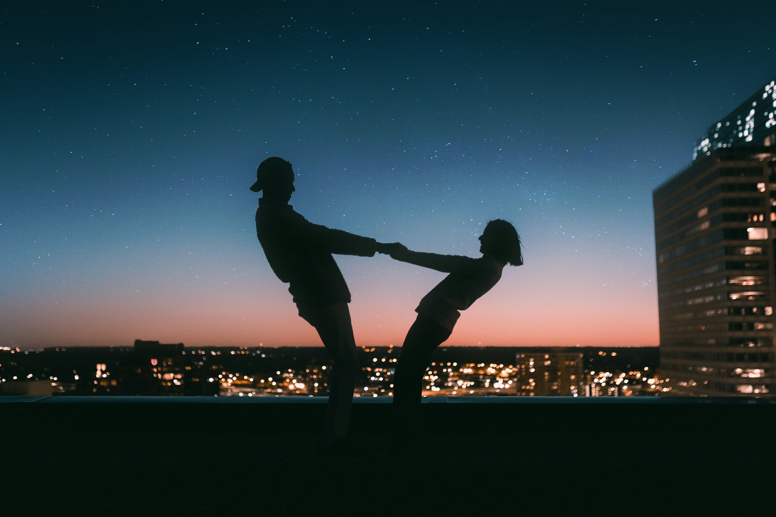 Silhouettes of two children holding hands and leaning back on a rooftop against a cityscape at sunset with a starry sky.