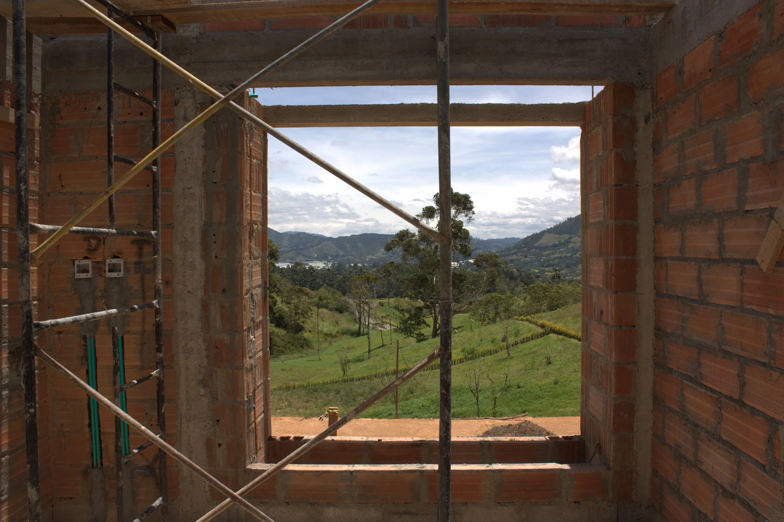 A small house on stilts with a wooden deck, lit from inside, set on a hillside surrounded by trees and mountains during dusk.