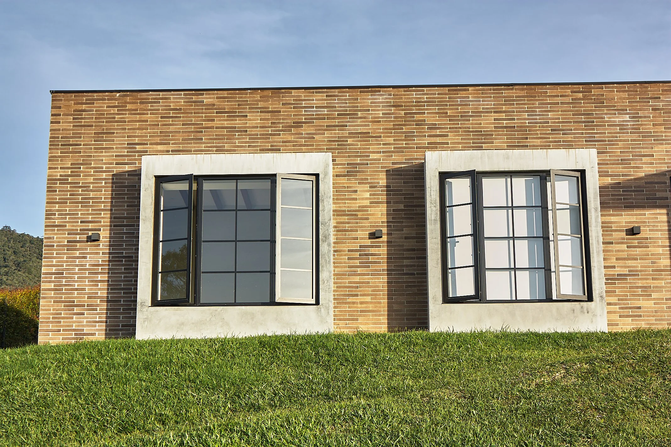 A brick building with two large, open, black-framed windows with white partial curtains behind glass, situated on a grassy hill against a blue sky.