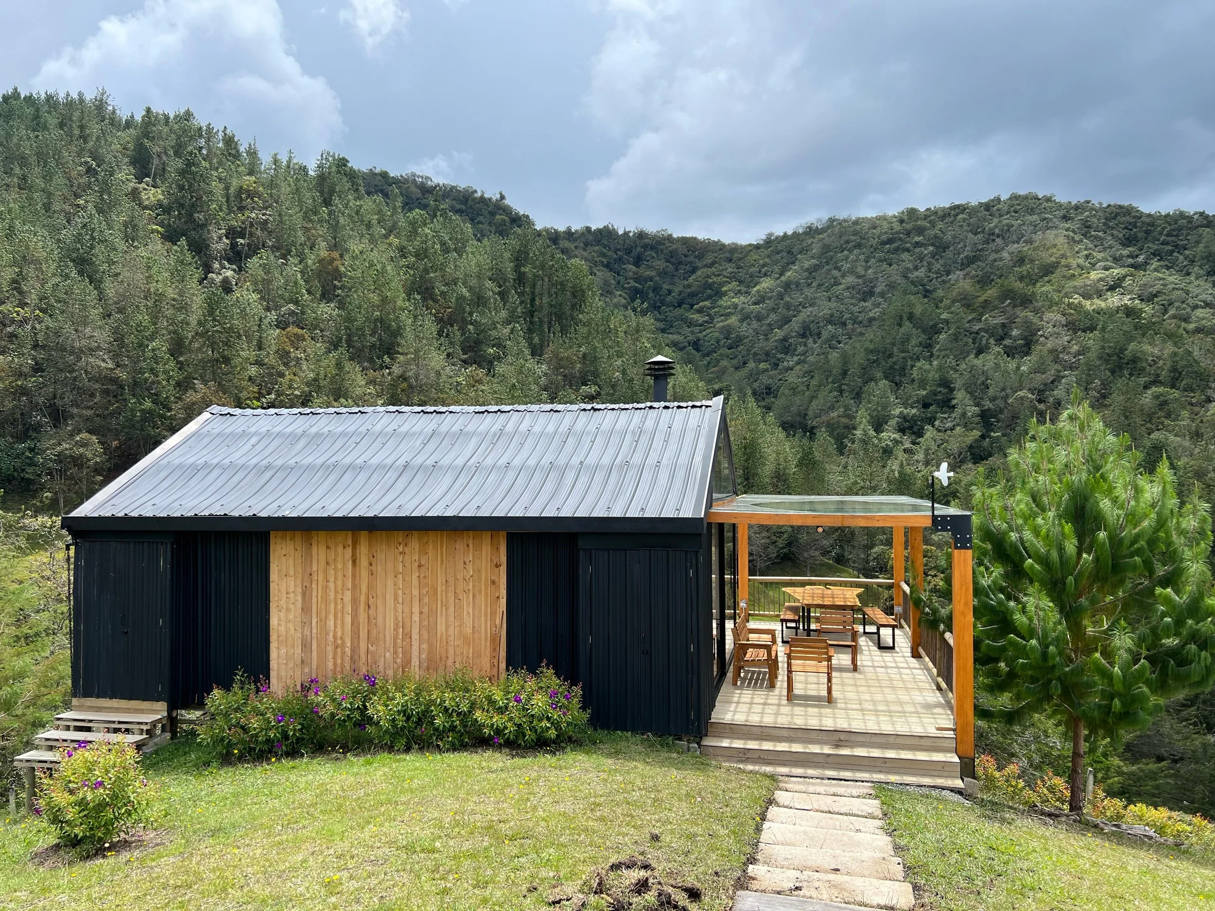 A small black and wooden cabin with a metal roof and a covered patio, situated in a lush green, hilly landscape.
