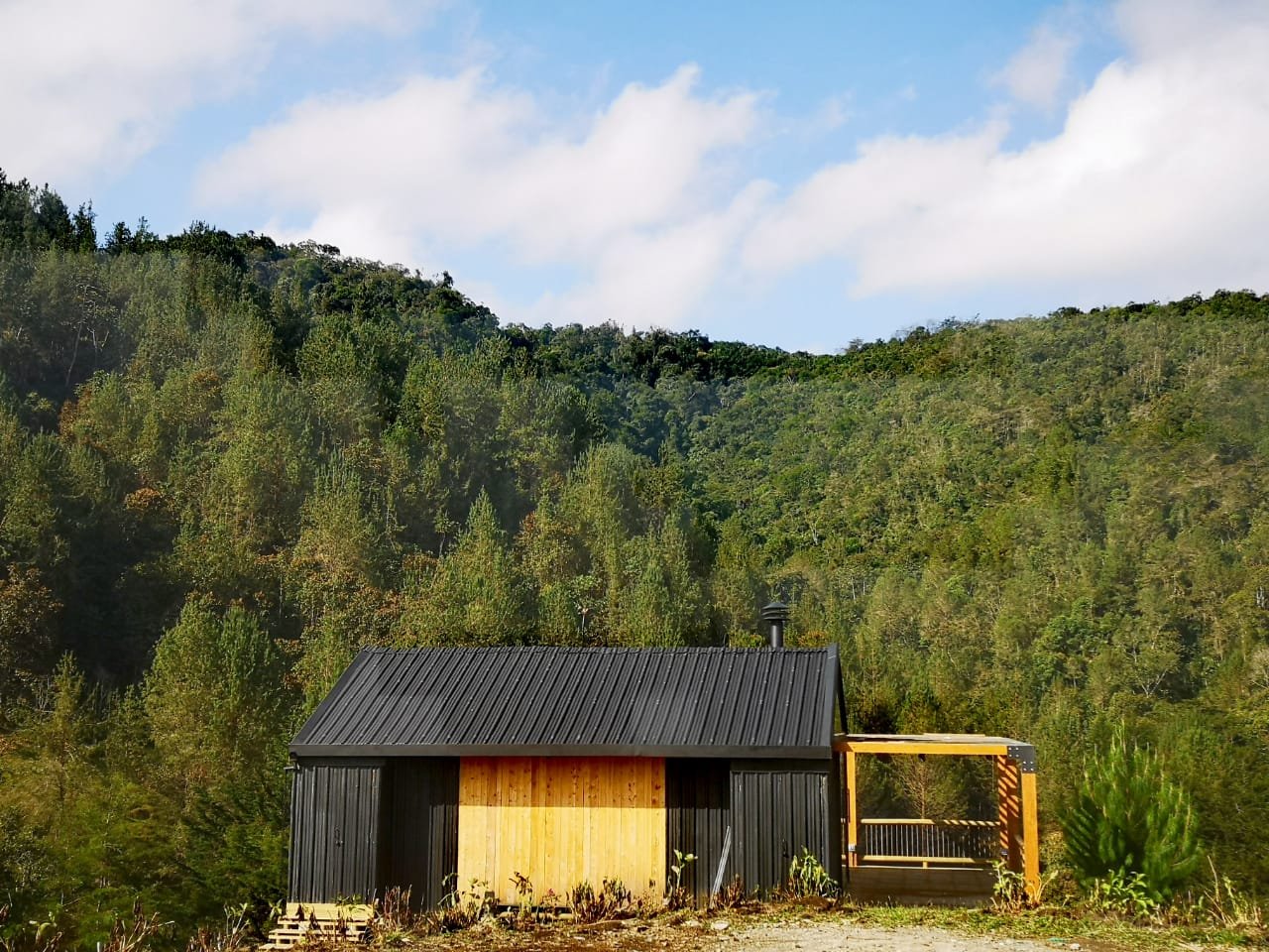 A small black and wooden cabin with a sloped metal roof, nestled in a green mountainous landscape under a partly cloudy sky.