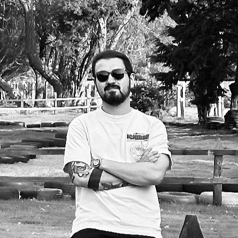 Man with sunglasses, short beard, tattooed arm, wearing a white t-shirt standing outdoors with trees and a wooden fence in the background