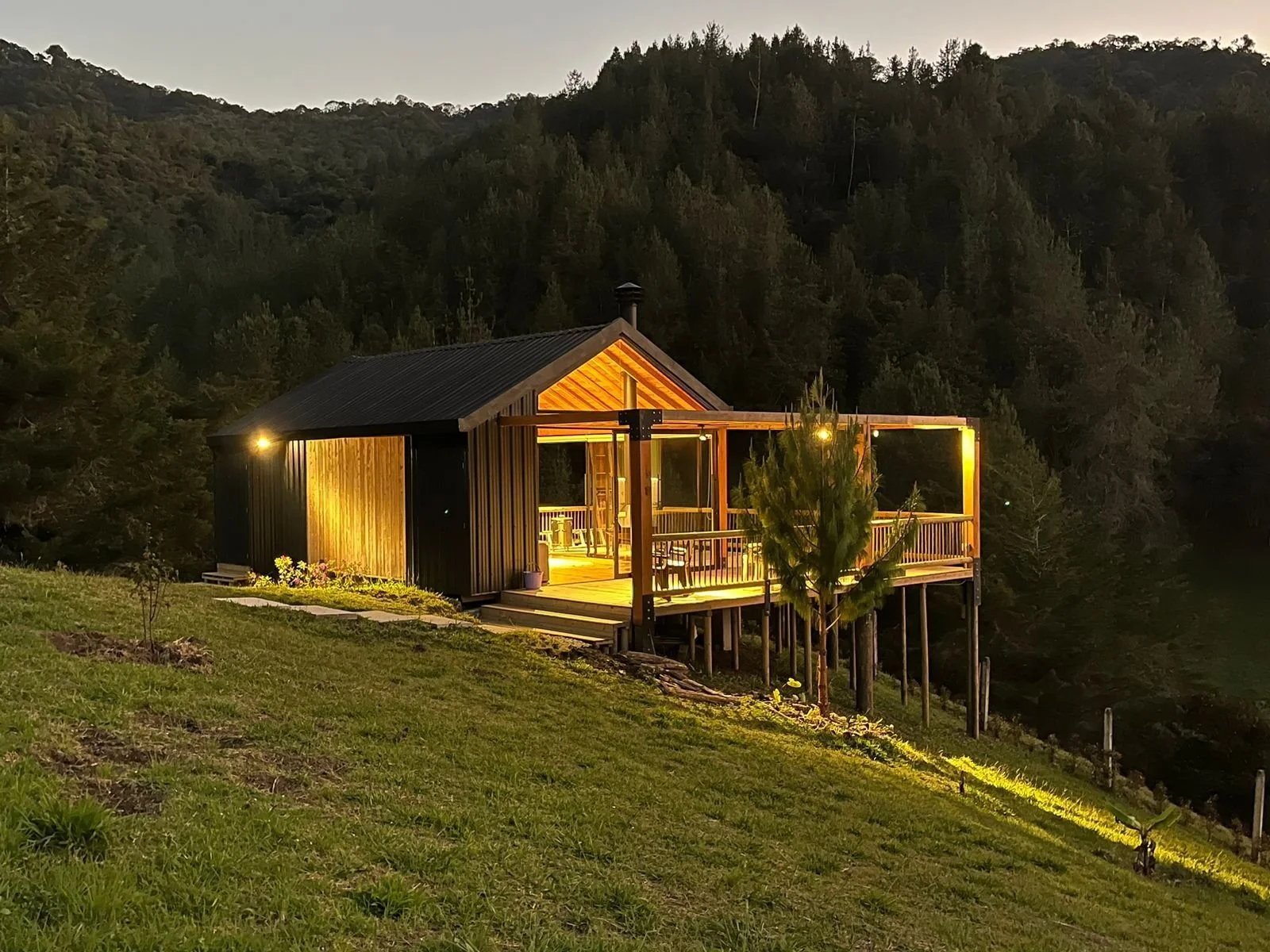 A small house with a black roof and wooden porch, illuminated at night, on a grassy hillside with trees and forested hills in the background.